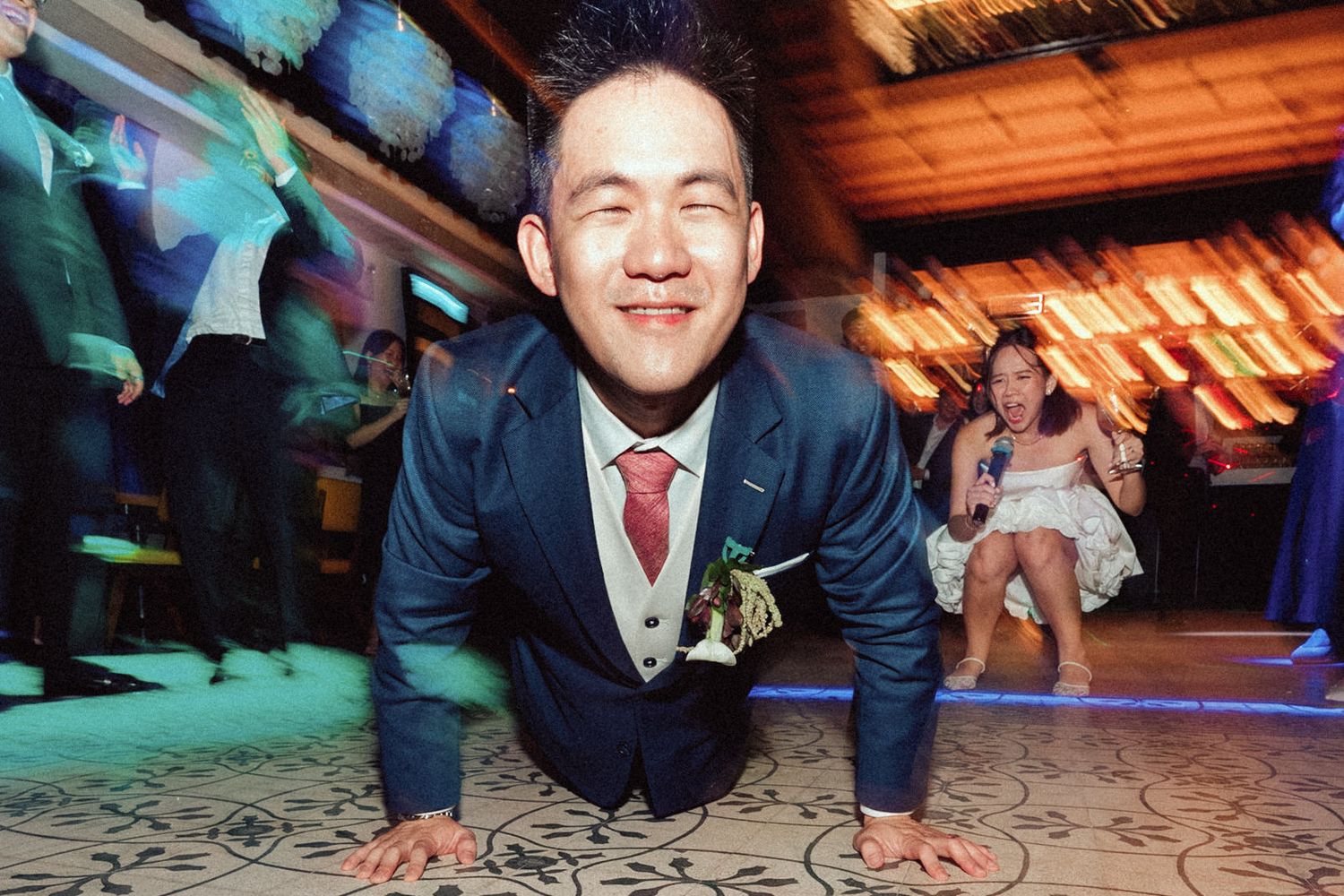 A wedding guest in a navy suit does a breakdance move on a patterned dance floor with colorful lighting in the background.