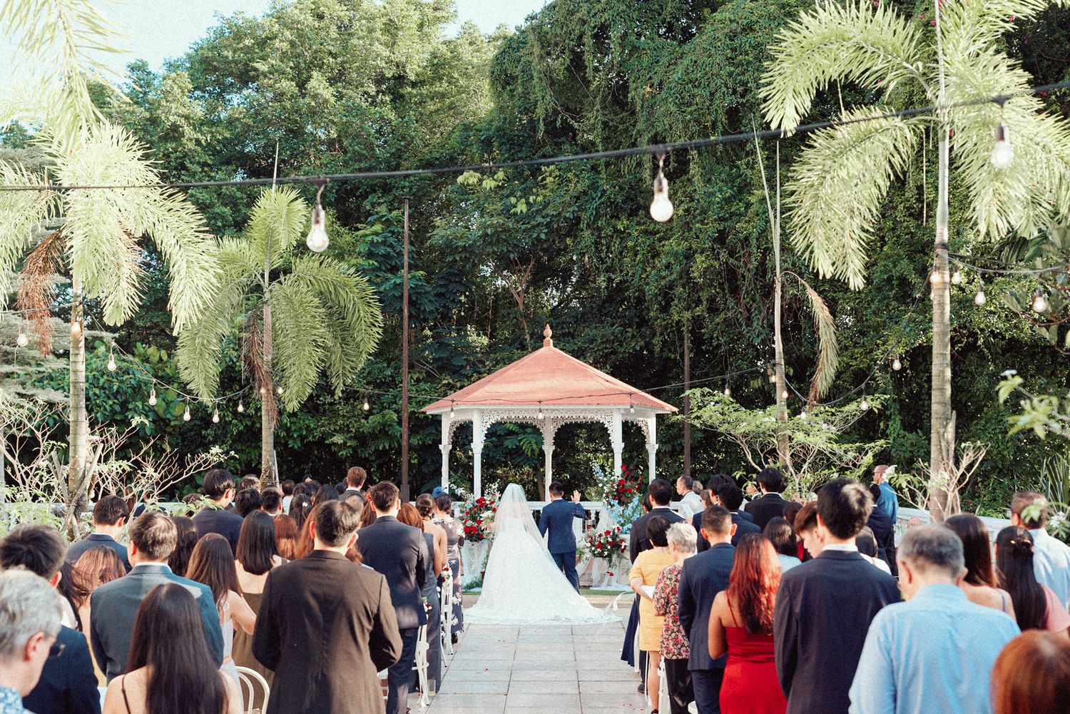Wedding guests gather outdoors under palm trees at a tropical venue with a white gazebo in the background.