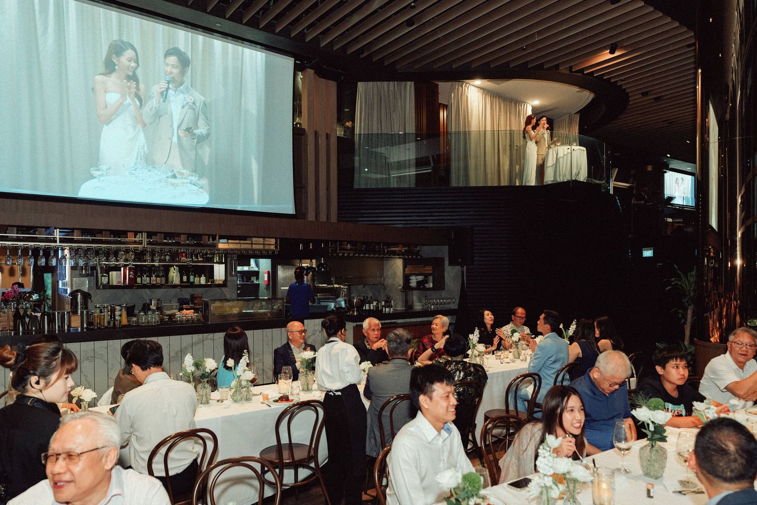 Wedding reception venue with large projection screen and guests seated at round tables.