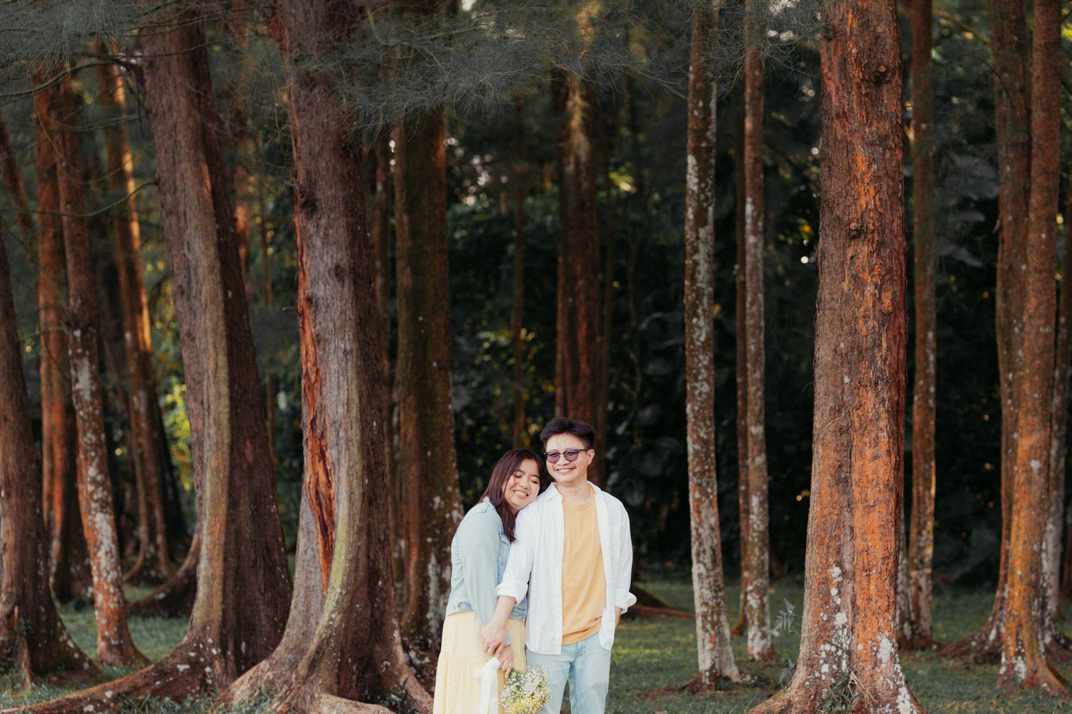 Couple embracing among tall pine trees in a lush forest, wearing casual outfits in a romantic outdoor photo session.