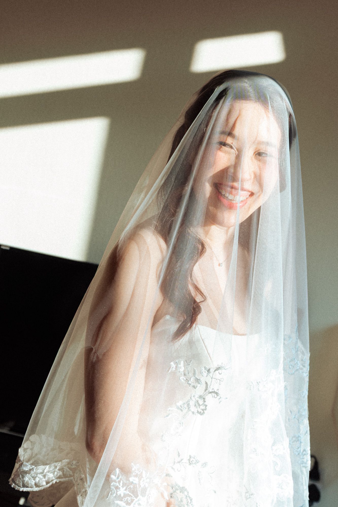 Smiling bride wearing a lace wedding dress and sheer veil draped over her face, standing in soft natural light.