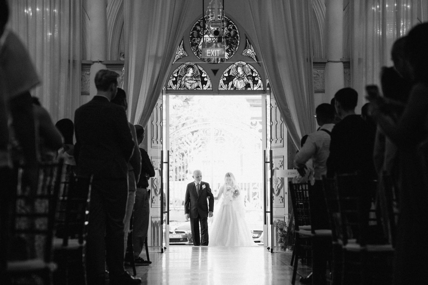 Black and white photo of bride and groom entering wedding ceremony through grand doors with guests seated in pews.