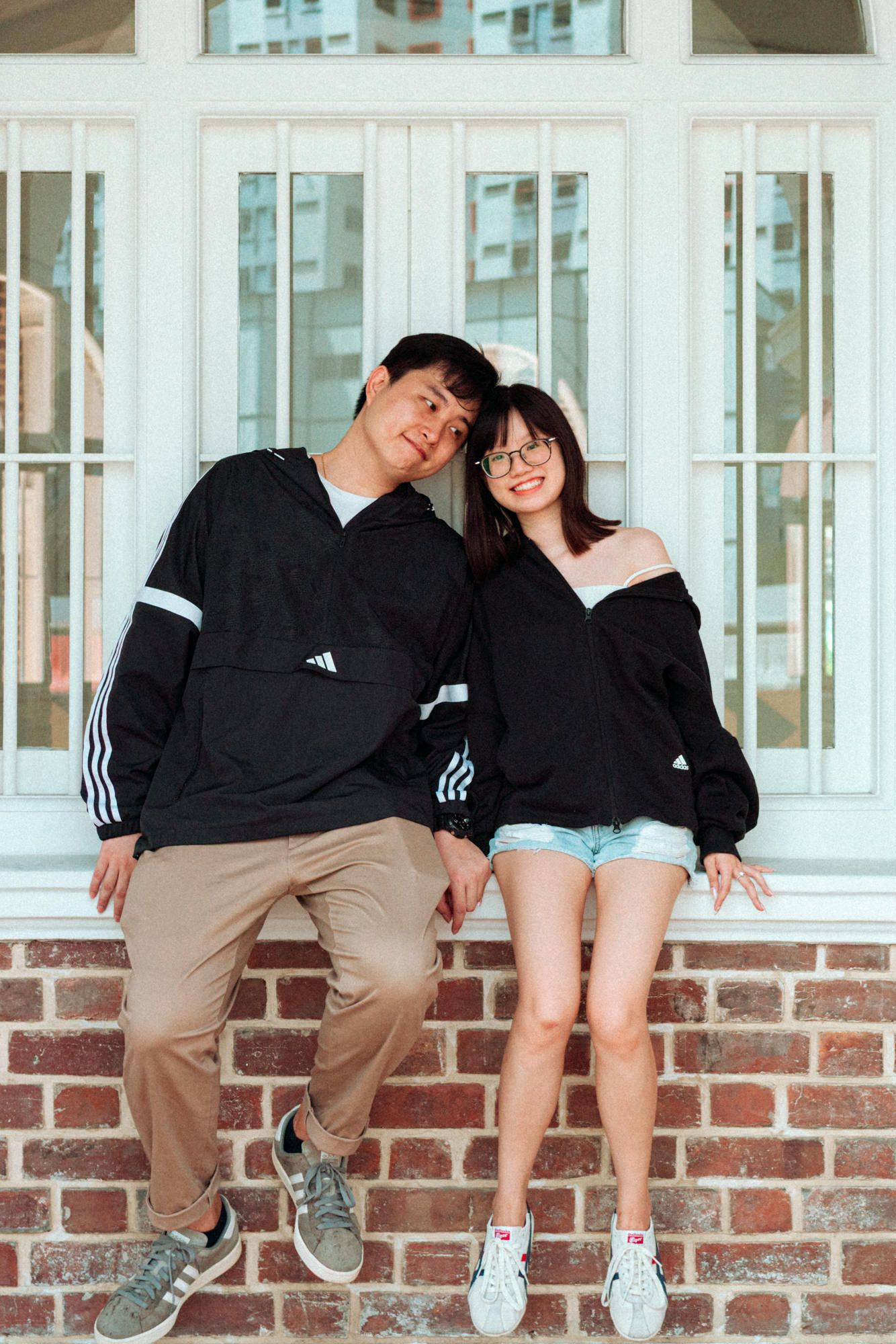 A couple in matching black hoodies and khaki pants sit together on a brick ledge in a romantic pose.