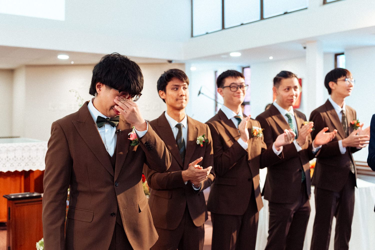 Emotional groom in brown suit with groomsmen clapping in church during wedding ceremony.