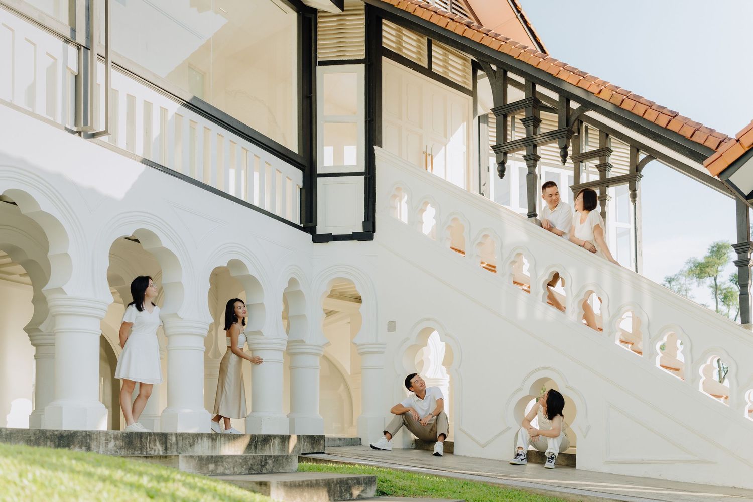People in white dresses pose on an elegant white colonial-style staircase with arched details and columns.