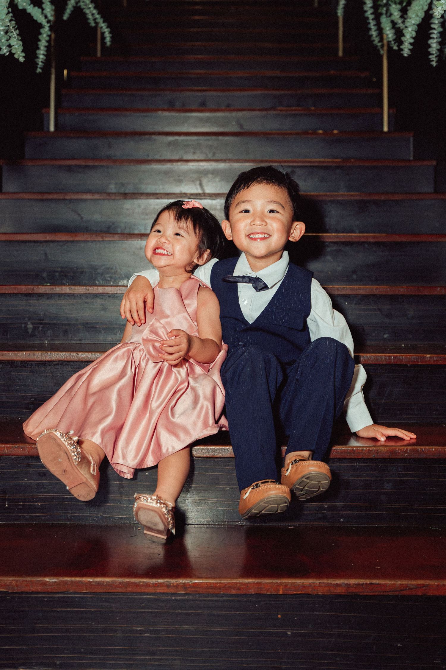 Two children in formal attire sit together on wooden stairs, wearing a pink dress and navy suit with bowtie.