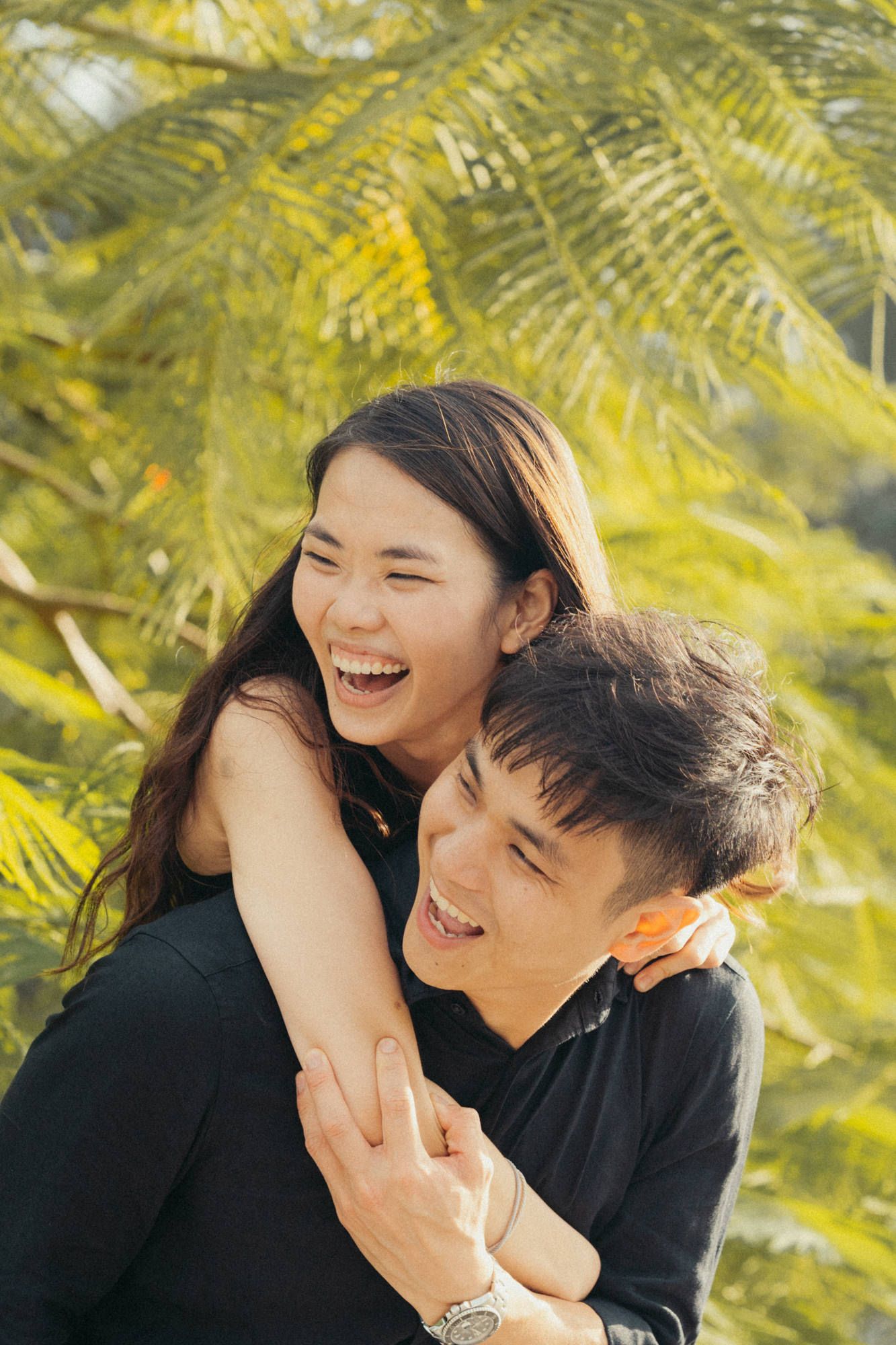 A romantic couple shares playful laughter together under palm fronds in natural sunlight.
