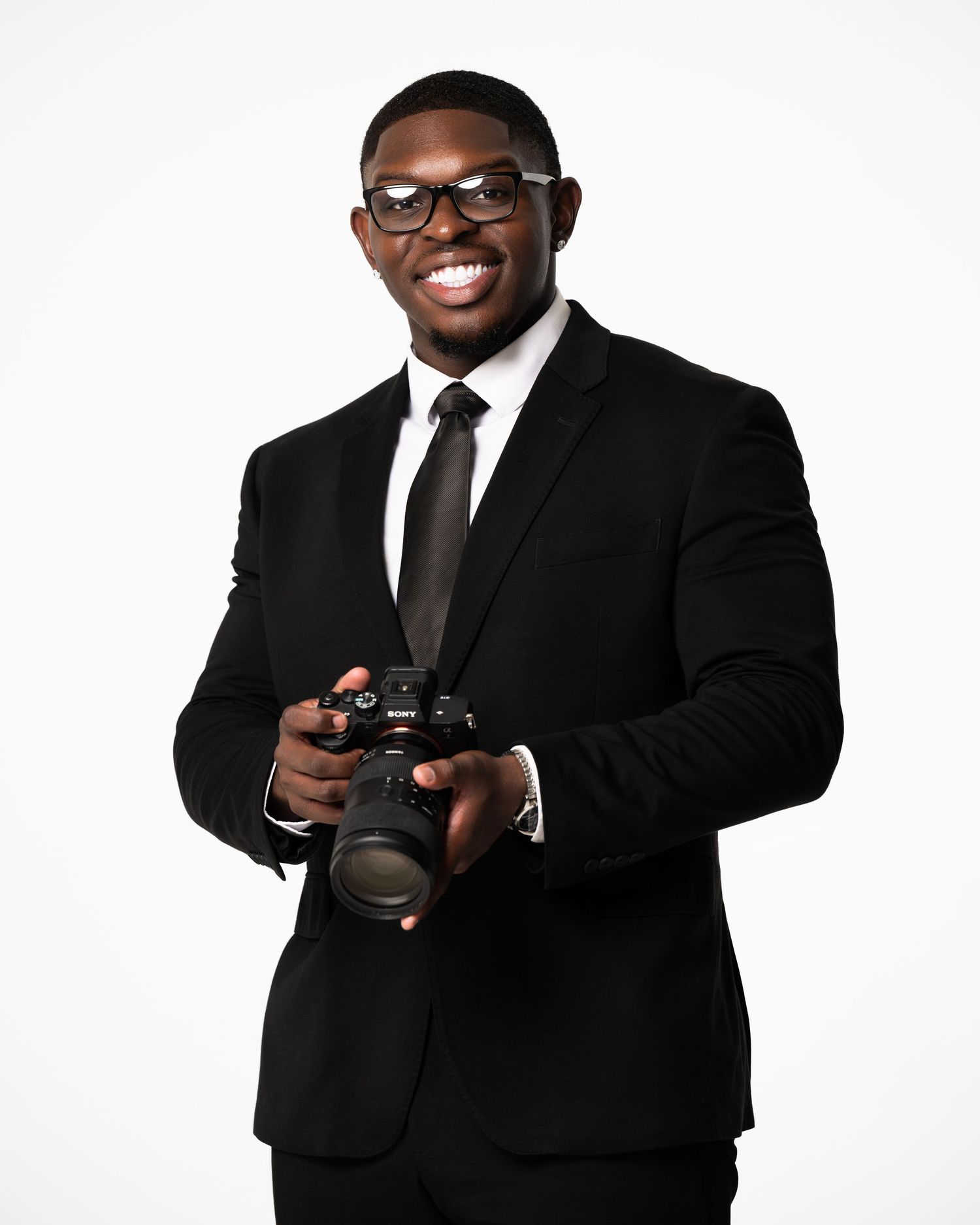 Professional photographer in black suit and glasses holds a DSLR camera while posing against white background.