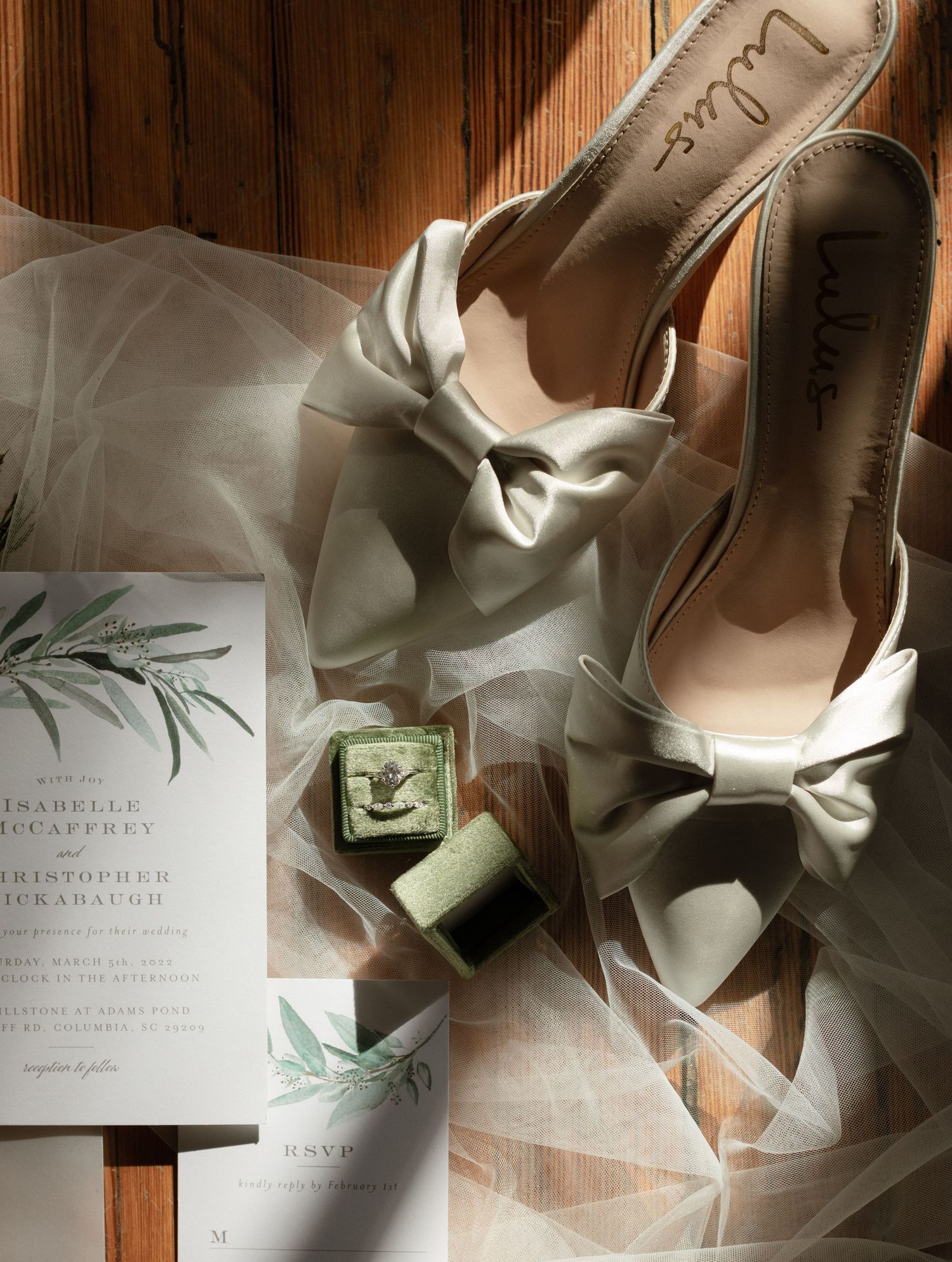 Wedding details including white heels and invitation suite arranged on wooden flooring with natural light.