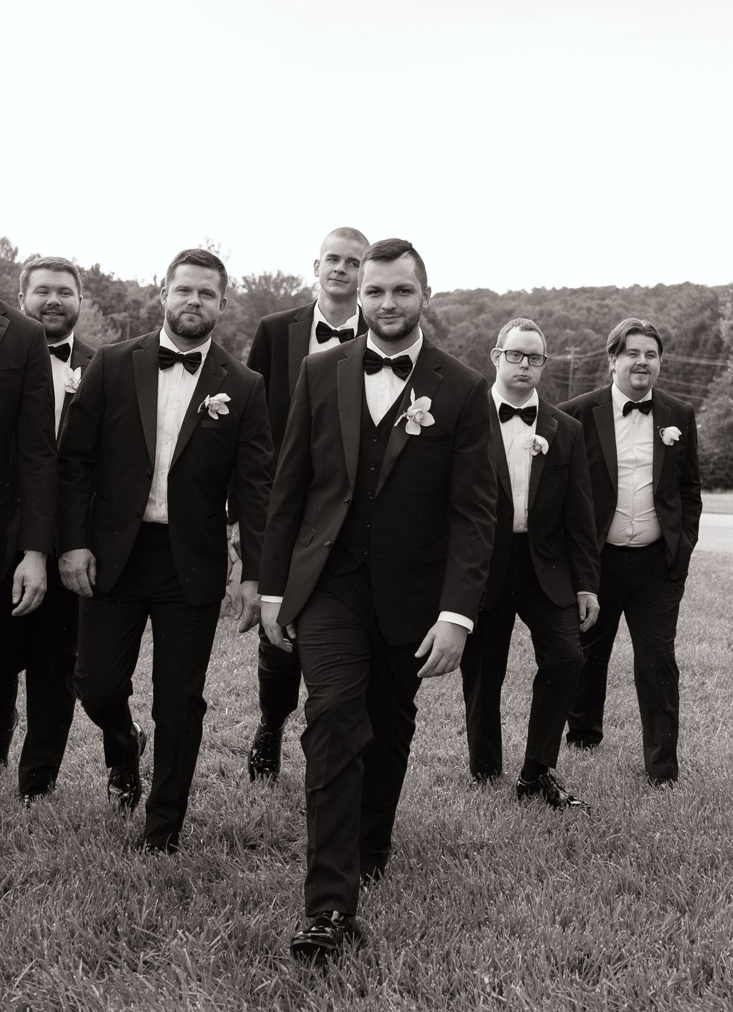 Groomsmen in black tuxedos pose creatively on grassy field.