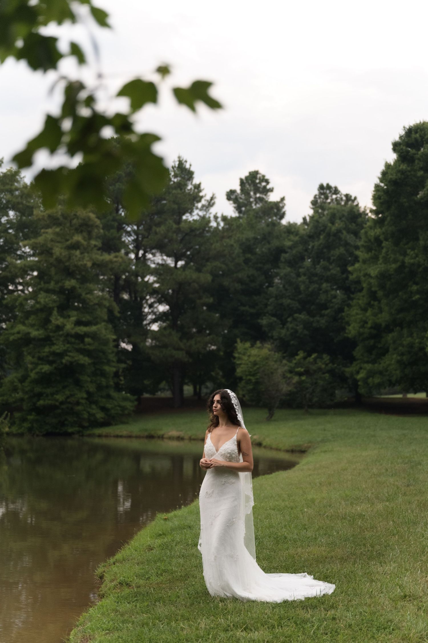 A bride in a flowing white gown stands near a peaceful pond surrounded by tall pine trees.