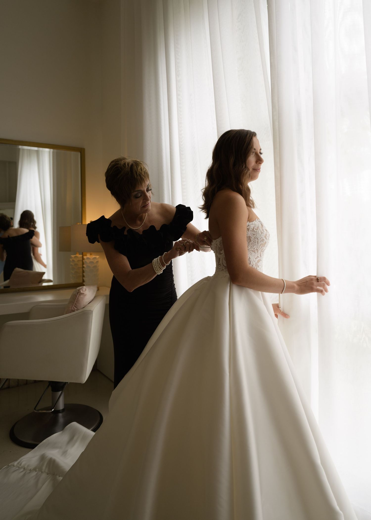 A white wedding gown being fitted and adjusted in a sunlit dressing room.