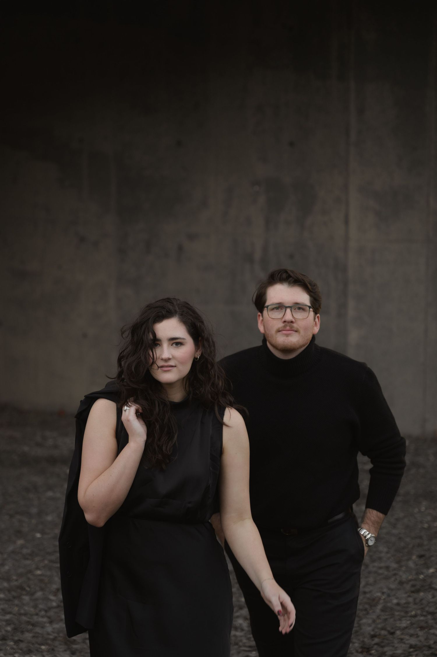 A couple dressed in black poses together against a dark backdrop in a moody dramatic portrait series.