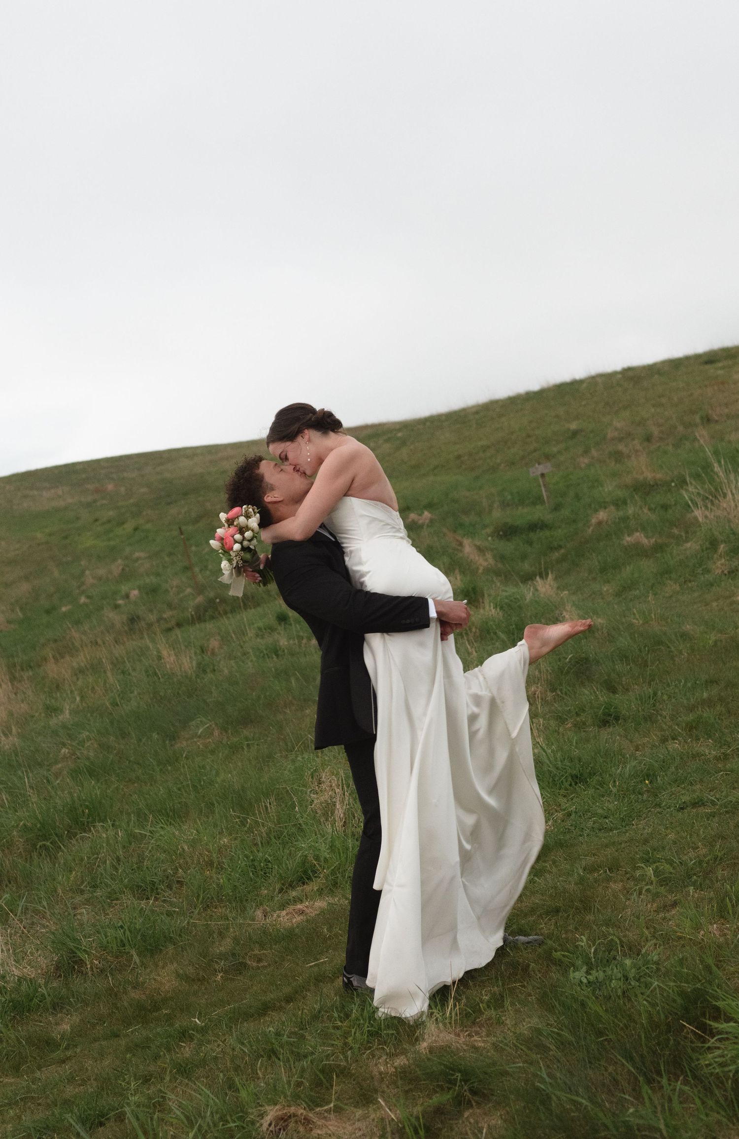 Wedding couple embracing on grassy hillside in dramatic pose.