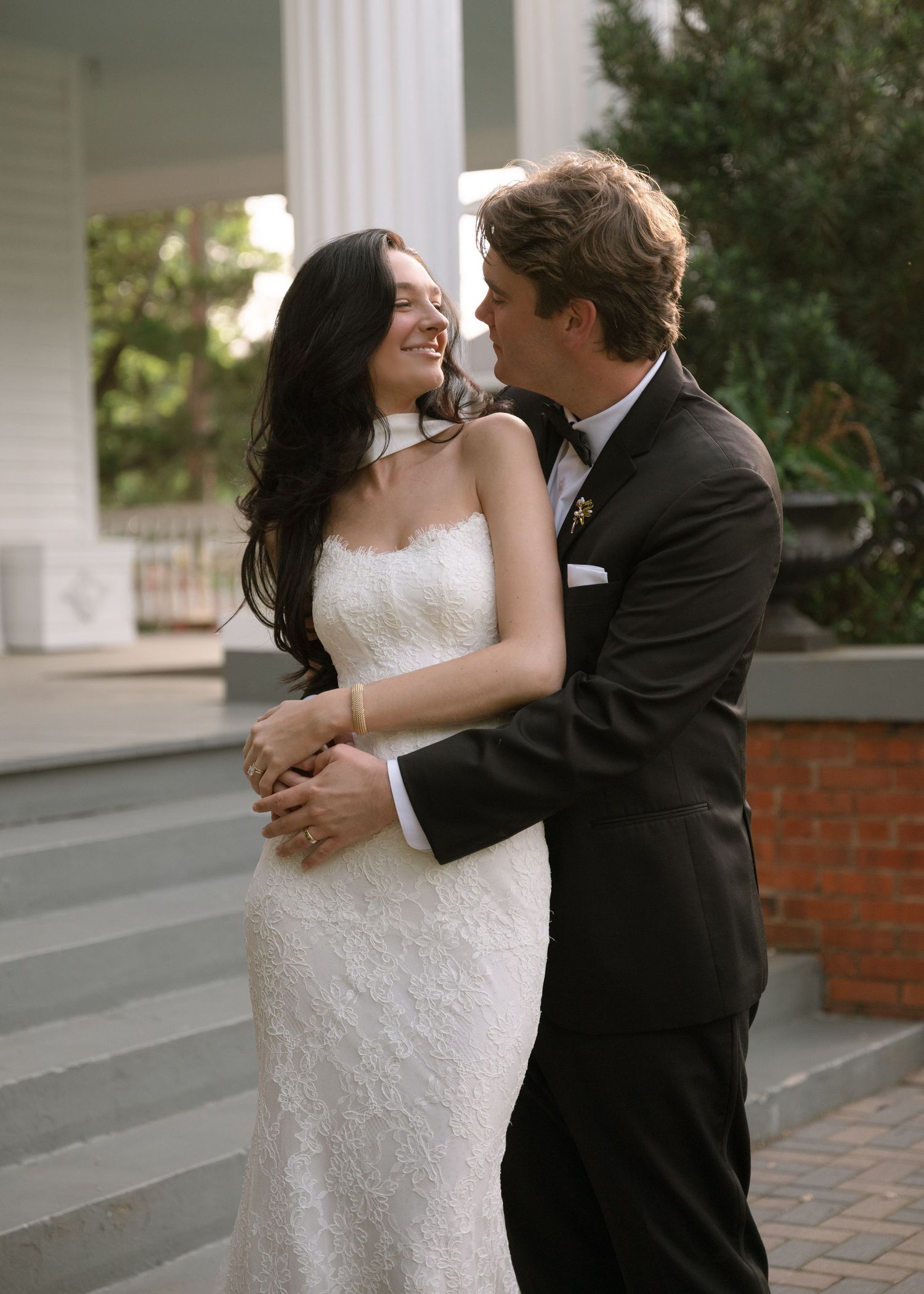 Bride and groom embrace outdoors near white columns, smiling at each other in wedding attire.