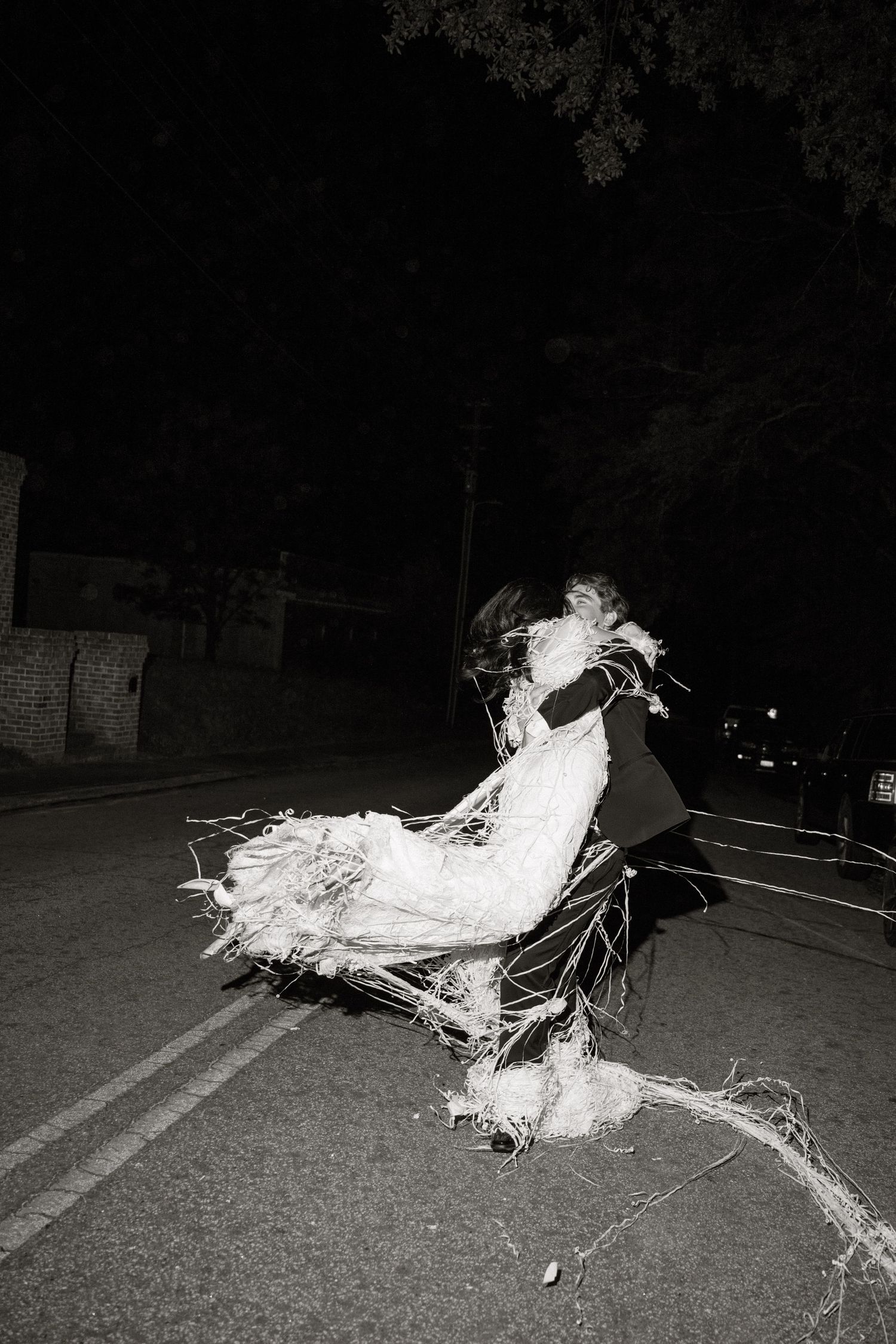 Black and white photo of a person in tattered white dress on dark street at night.