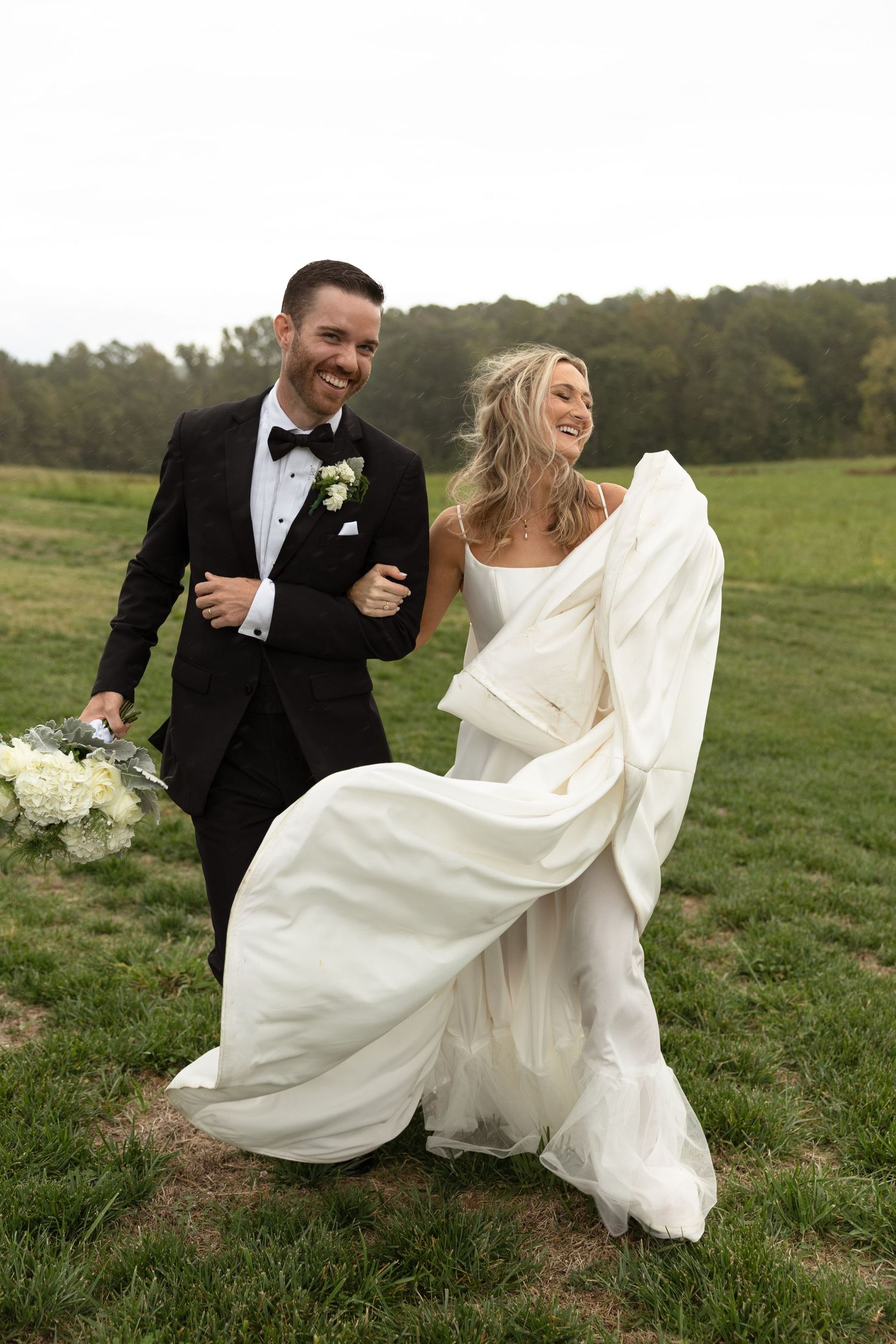 A couple laughing together in a grassy field wearing formal wedding attire.