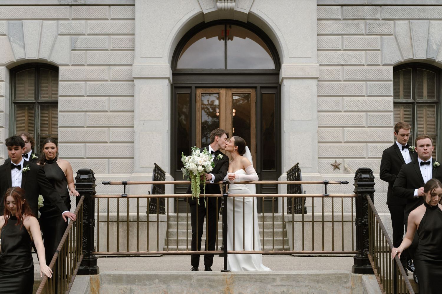 A group of people stand outside a stone building with arched doorways and windows during a formal gathering.
