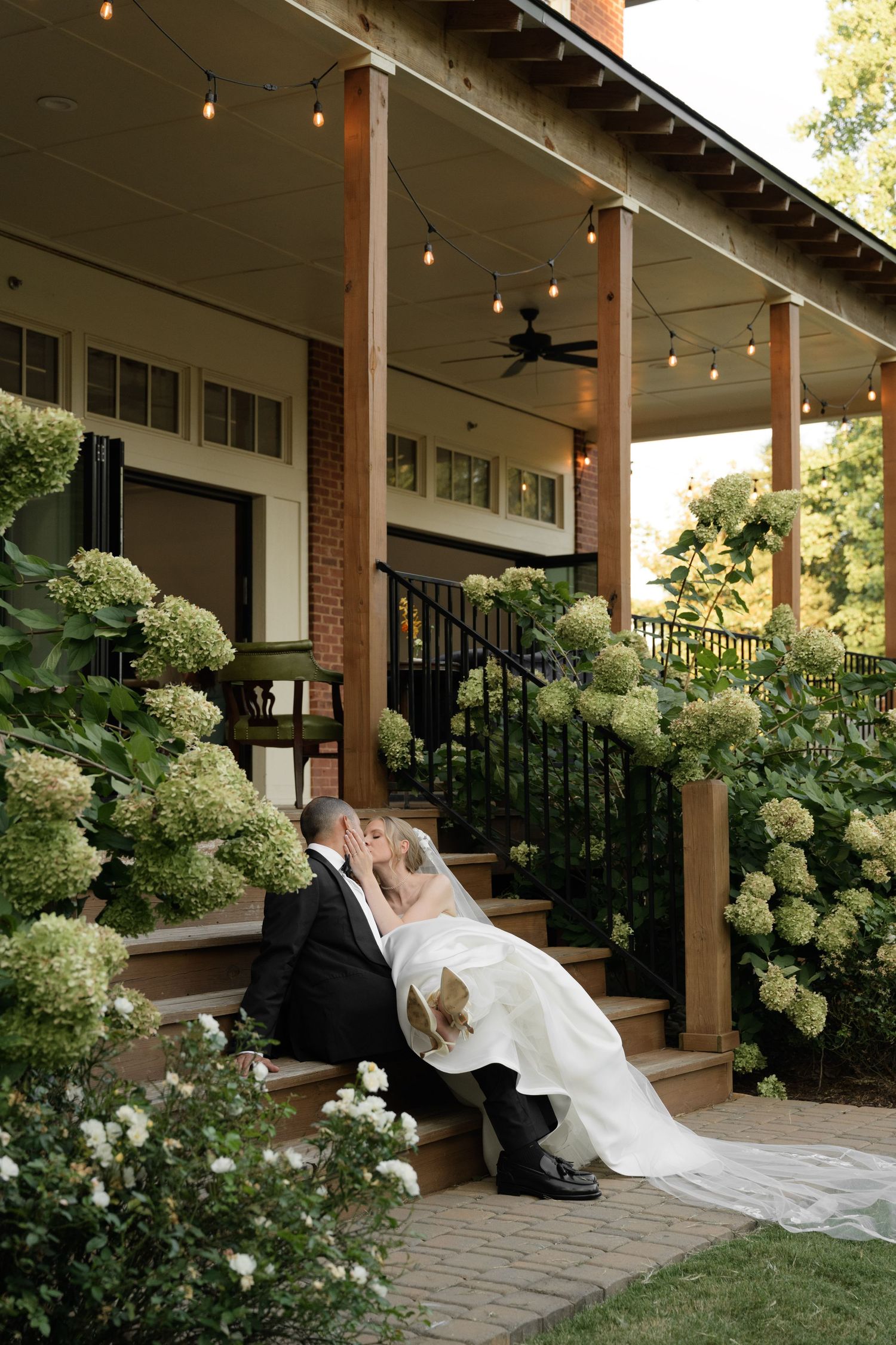 Newlyweds share a romantic moment on a porch decorated with white hydrangeas and string lights.