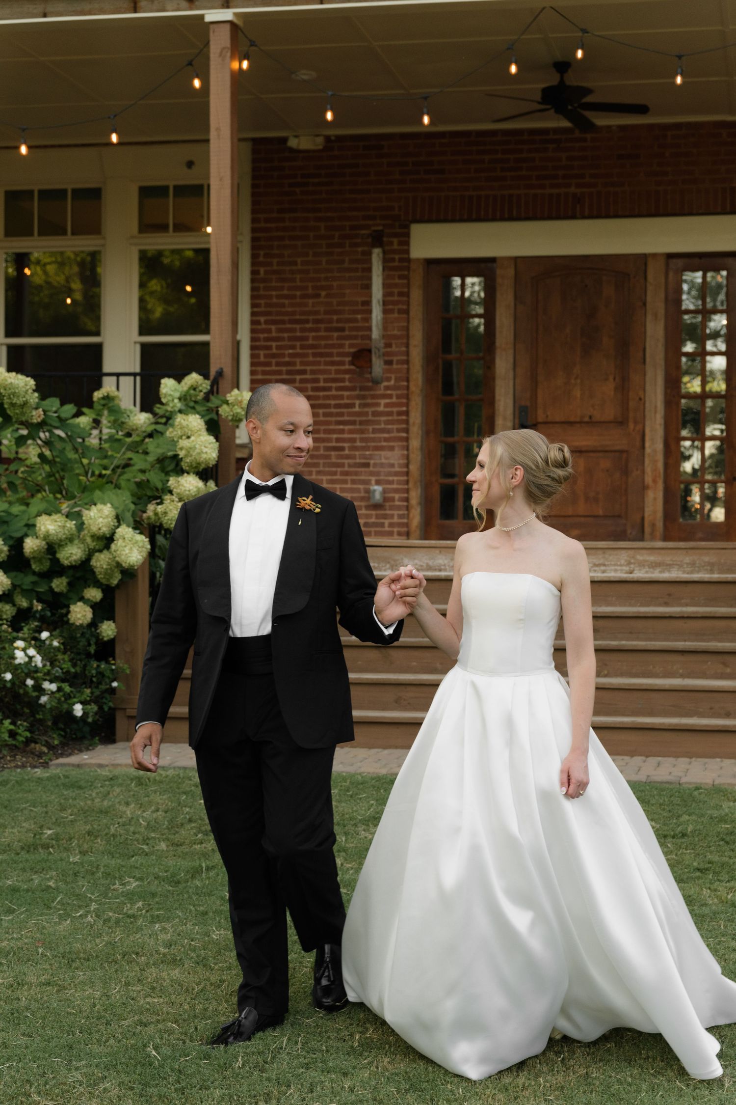Couple in formal wedding attire standing on lawn in front of brick house with string lights.