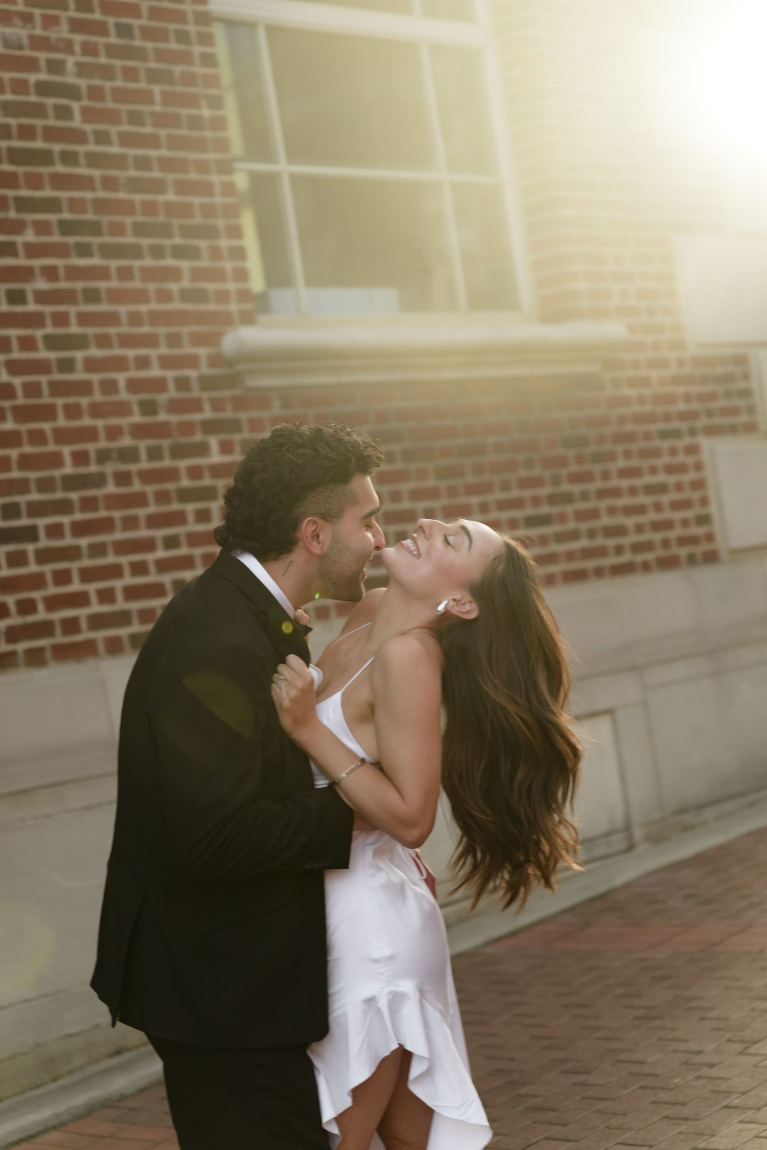Dancing couple in formal attire share playful moments against a brick wall.