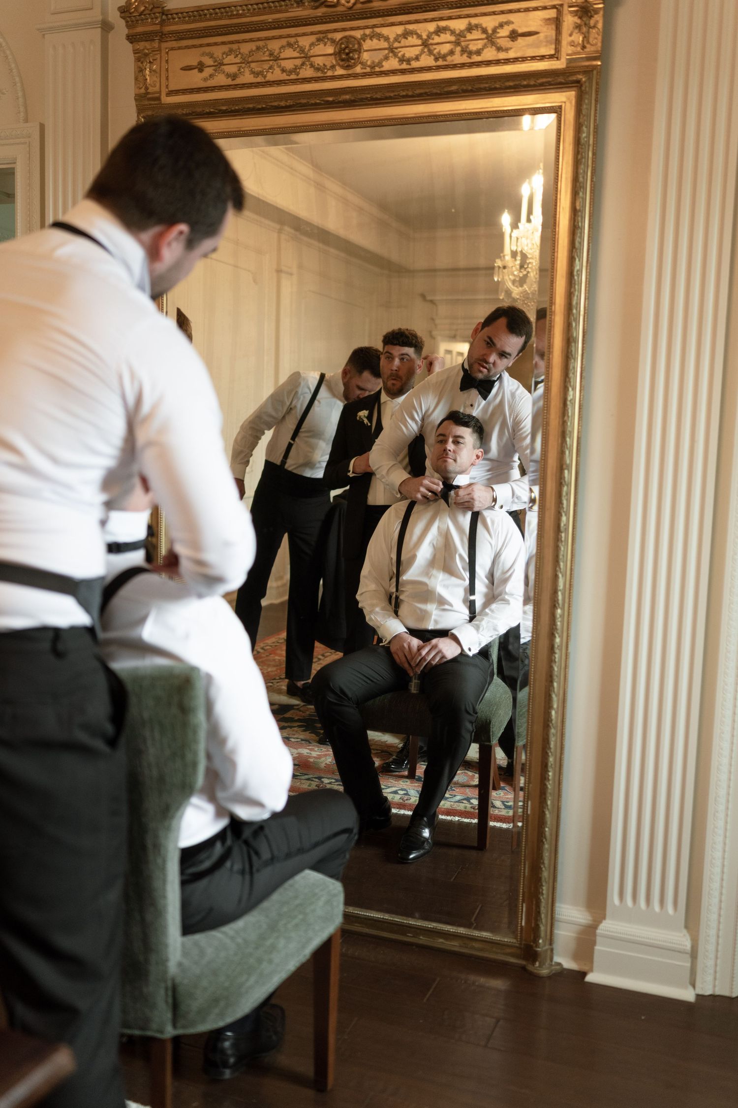 Wedding party getting ready in front of ornate gold mirror in formal attire.