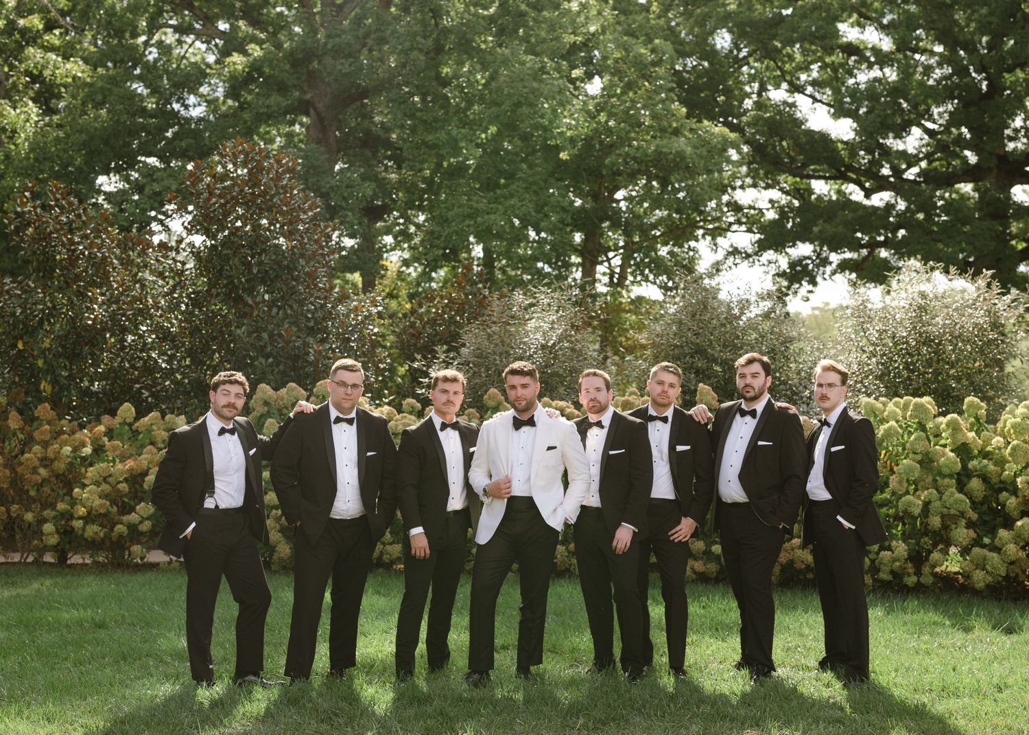 Groomsmen in black suits and ties stand in line against a lush green outdoor backdrop during a wedding photo session.