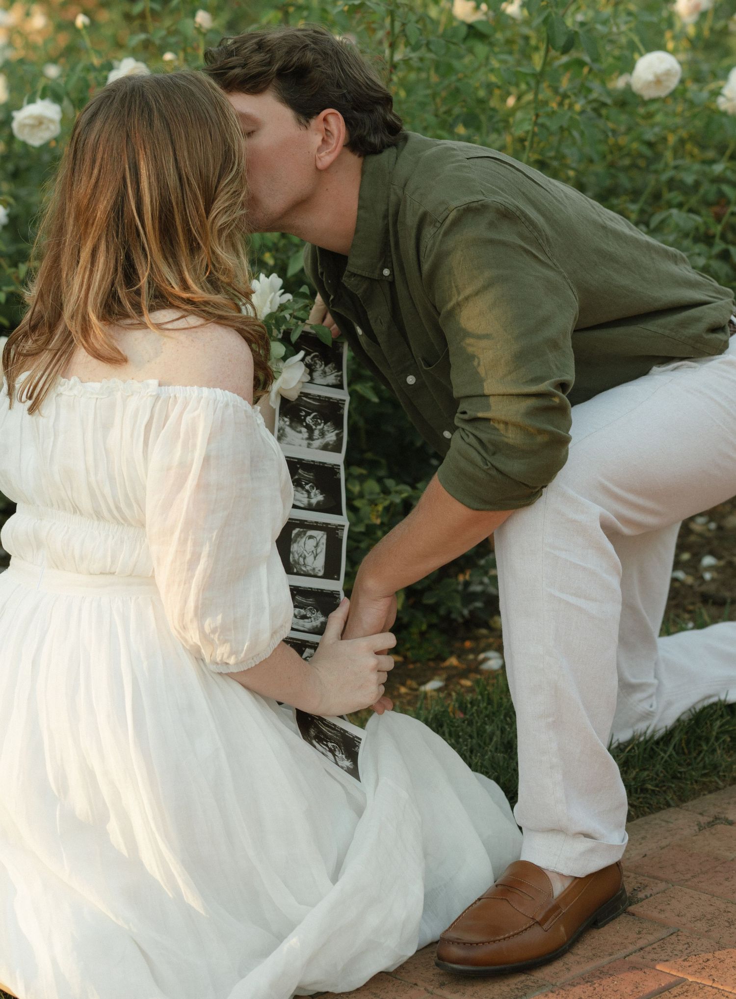 Couple in wedding attire sitting together in a garden with white roses.