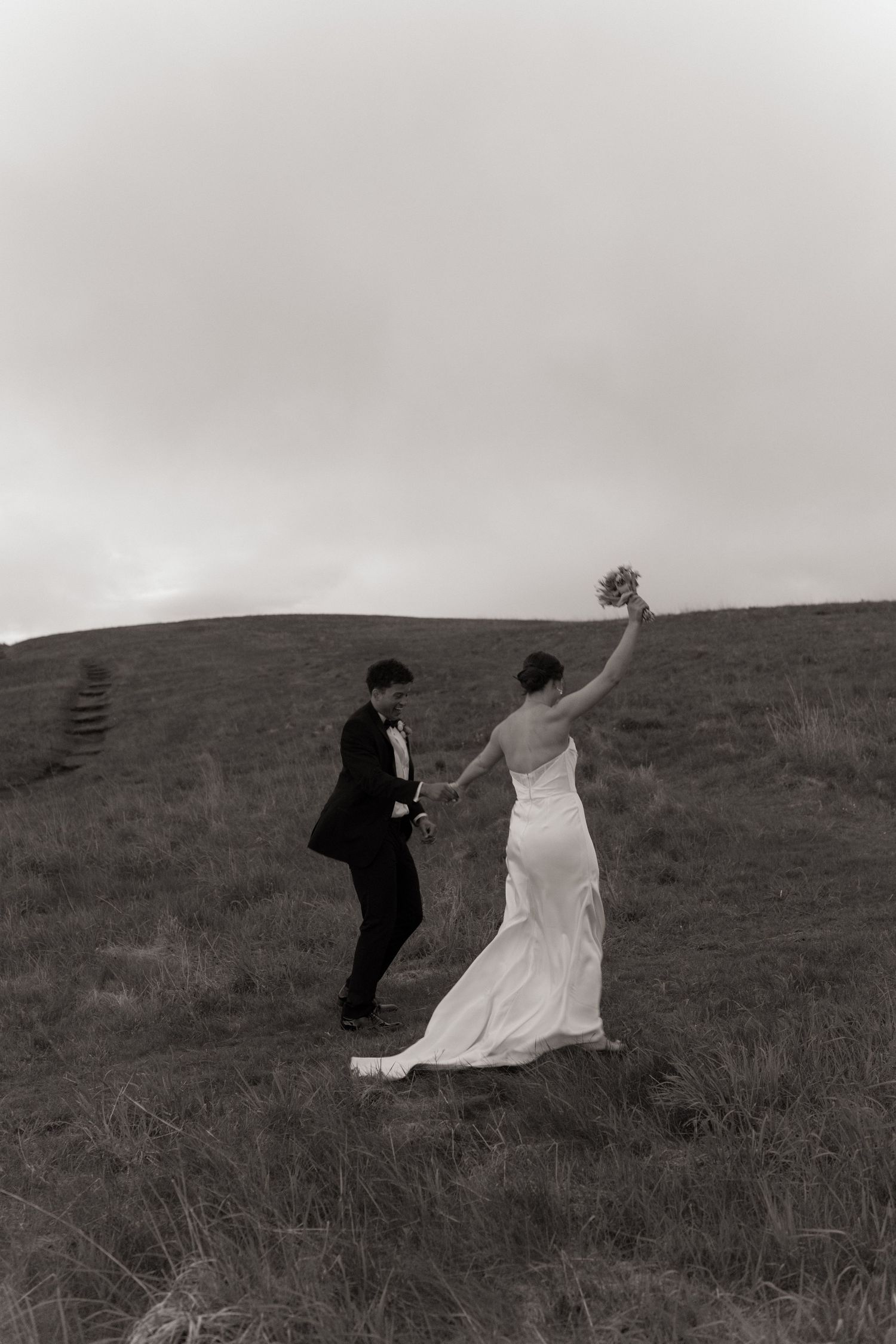 Black and white photo of couple in wedding attire playfully running across a hilltop.