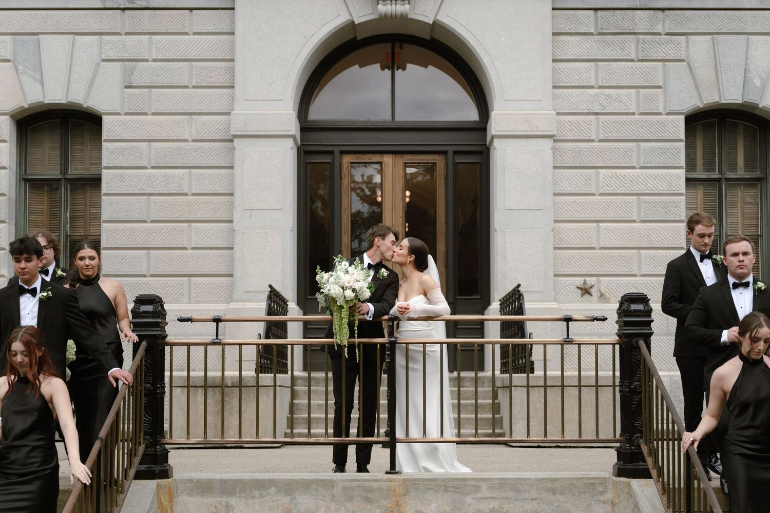 Wedding party members gather on stone steps outside a historic building entrance for formal photographs.