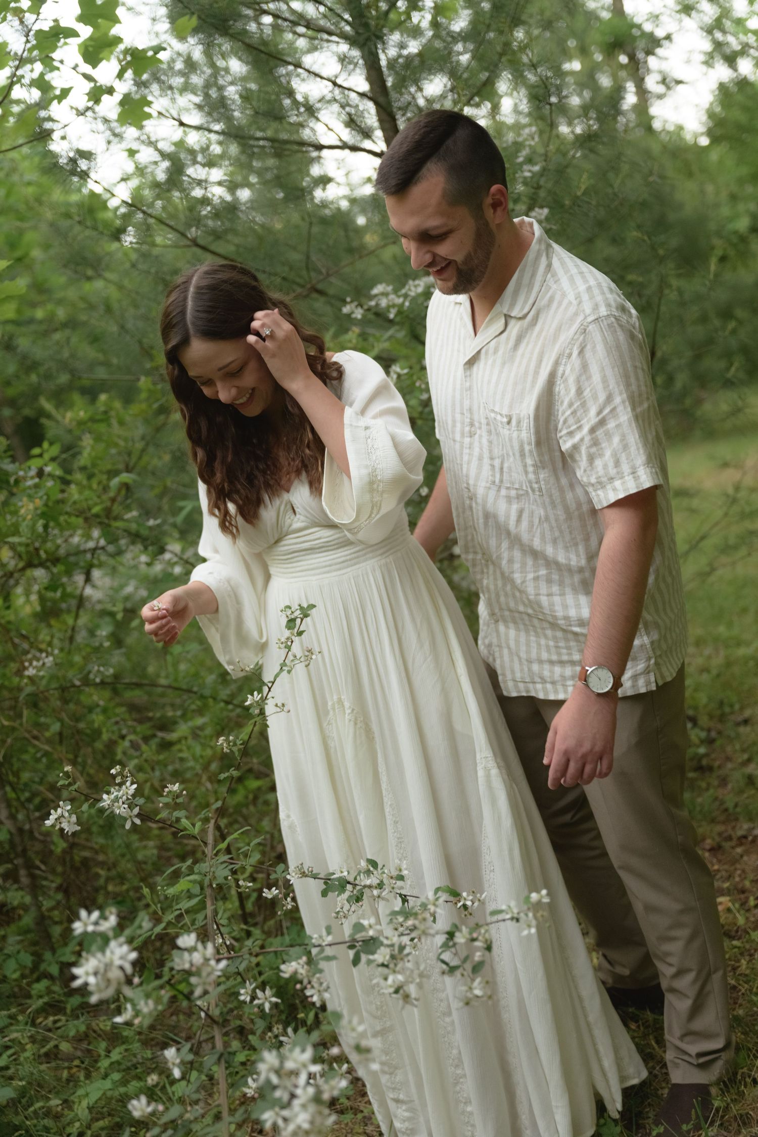 Romantic couple photoshoot in white attire among green foliage and trees.
