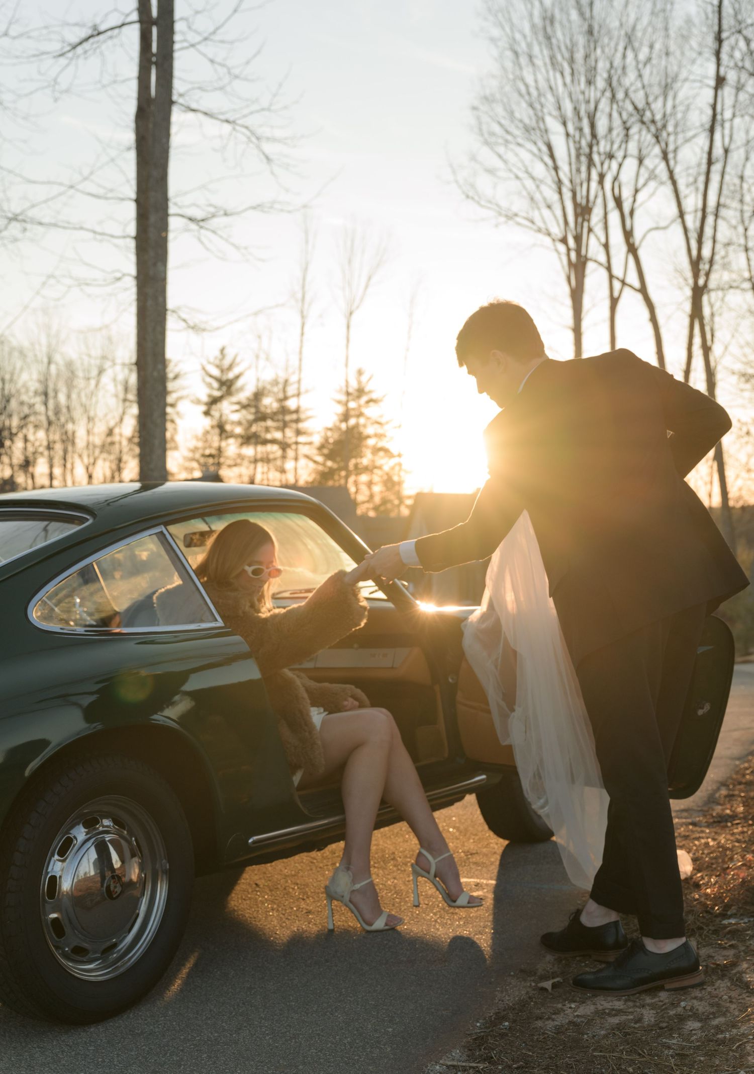 Silhouetted wedding couple near a vintage car at sunset with dramatic lighting.