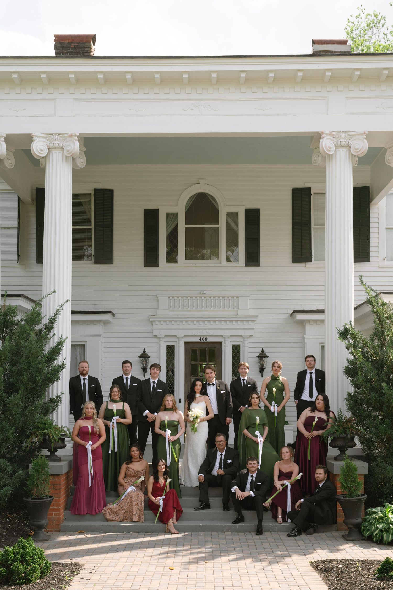 Wedding party posing in front of a white colonial mansion with columns, bridesmaids in green and red gowns, groomsmen in black tuxedos.