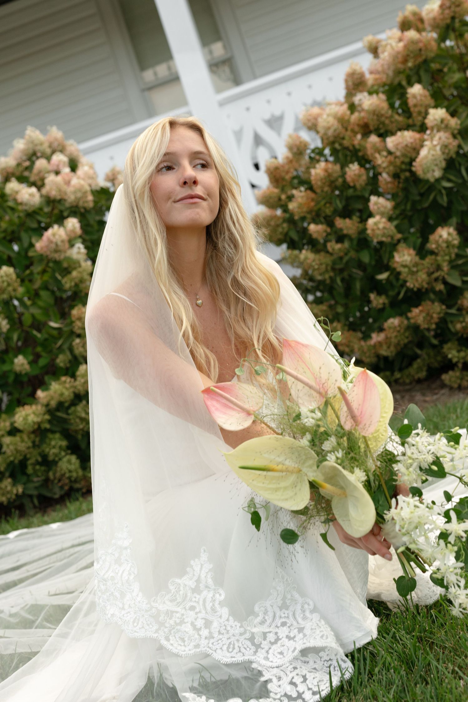 Bride in lace wedding dress poses with bouquet against blooming hydrangea bush.
