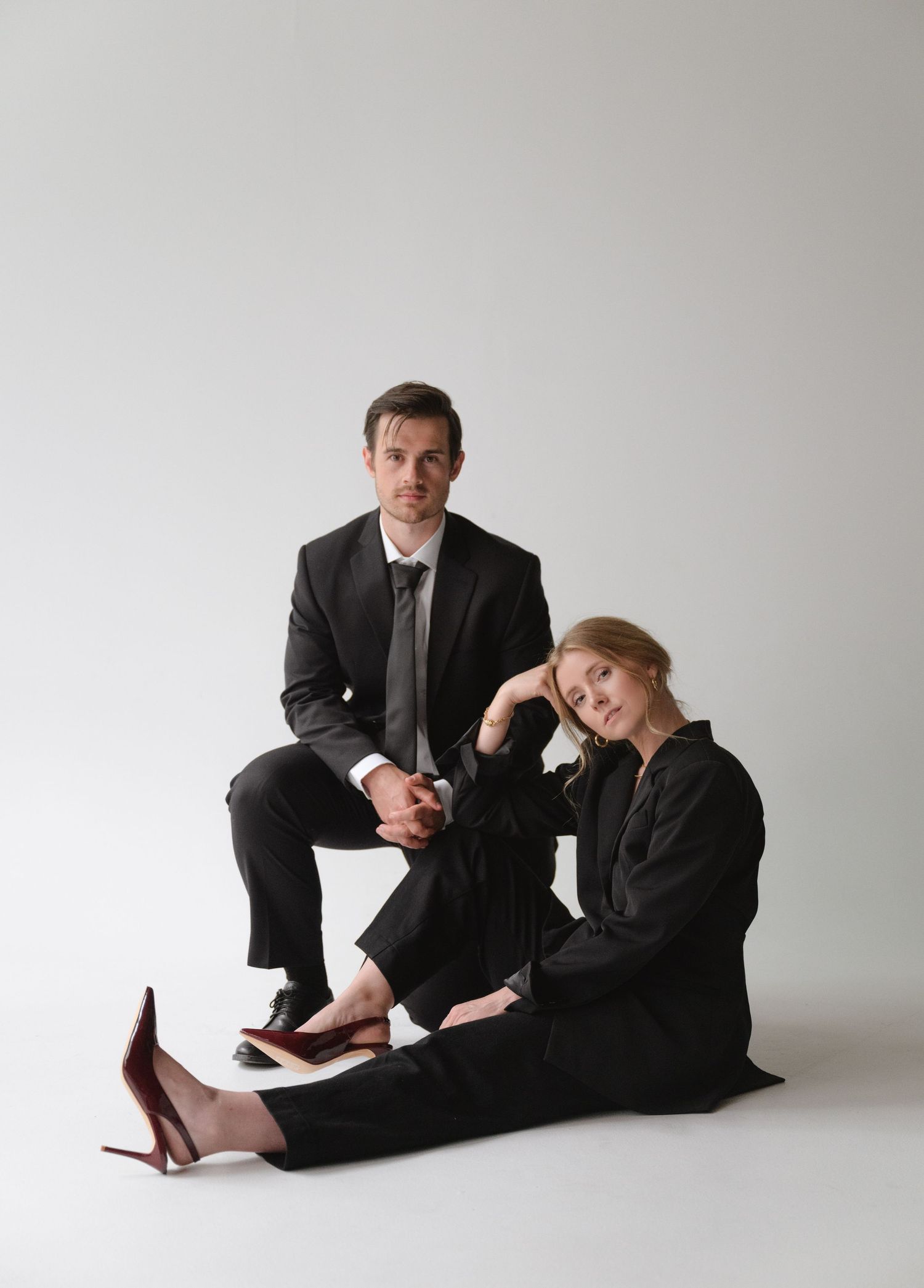 Professional photoshoot featuring two people in black suits on a minimalist white background.
