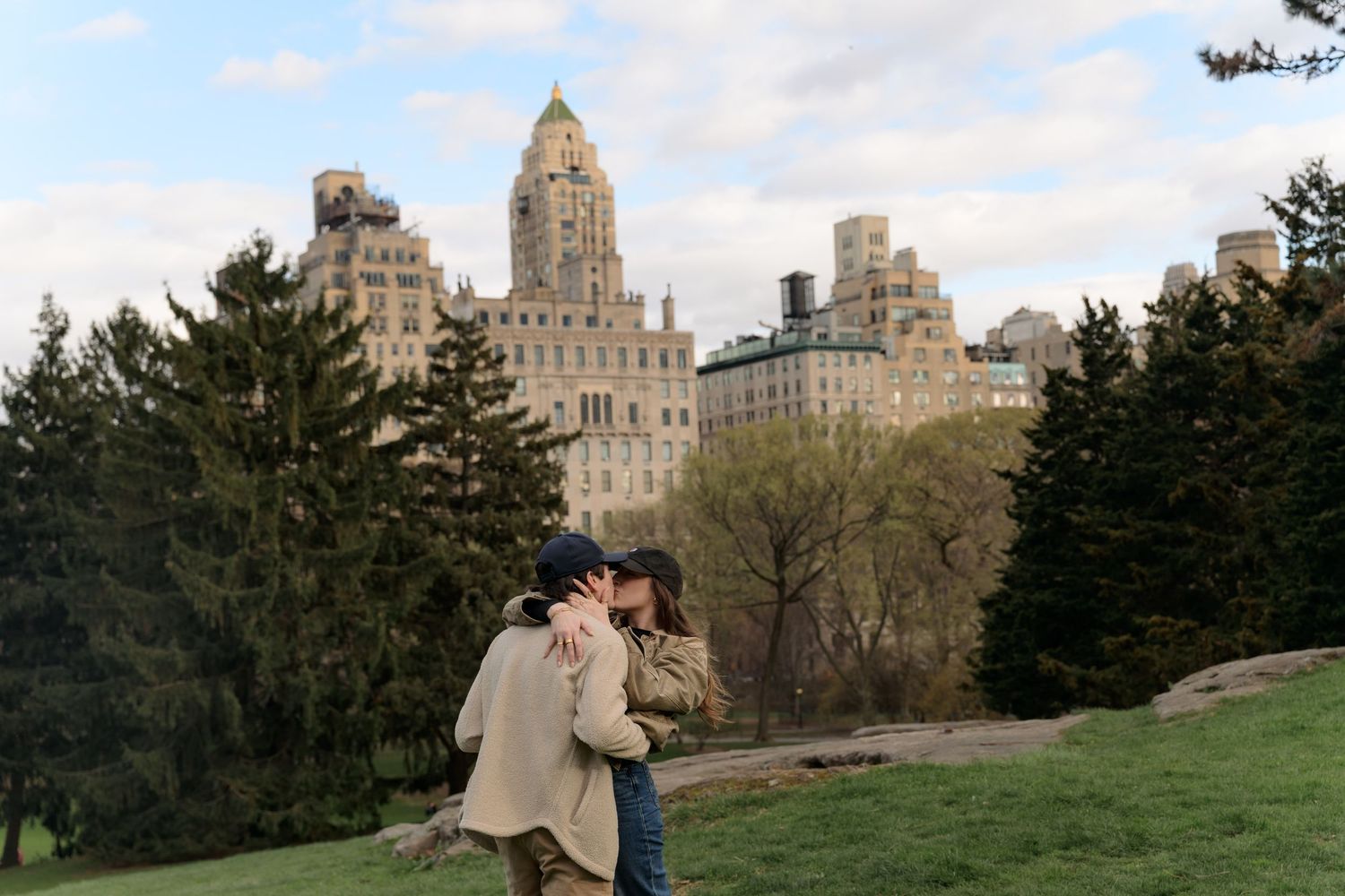 Romantic proposal sequence photographed in a grassy park with city buildings in the distance.