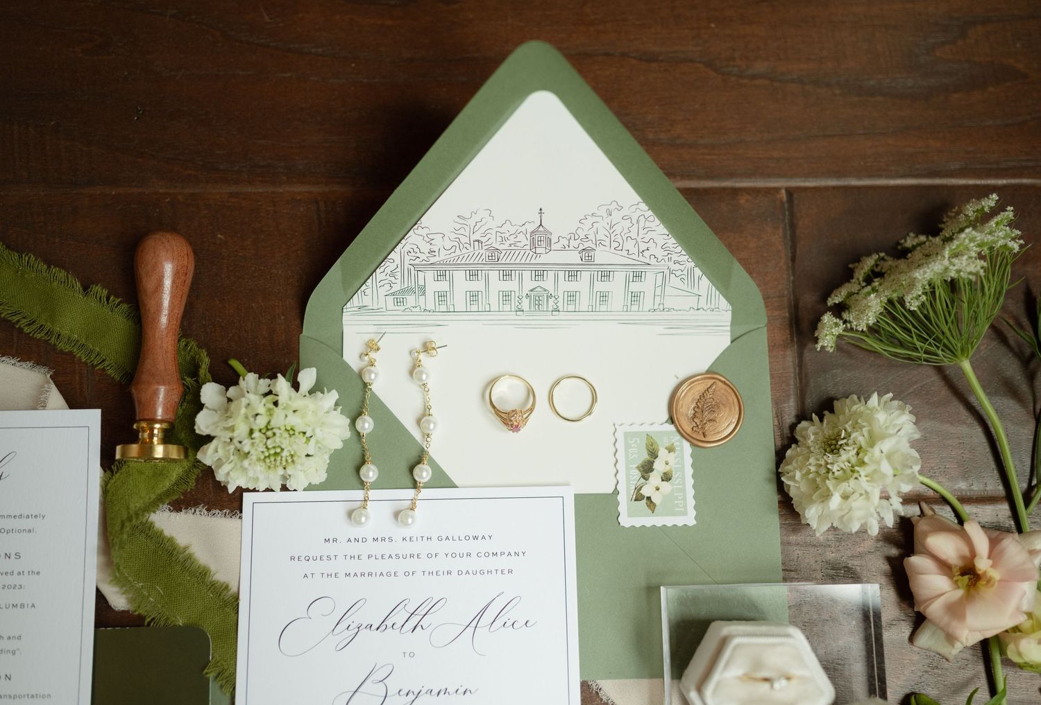 Wedding stationery flat lay with green envelope, rings, and white floral accents.