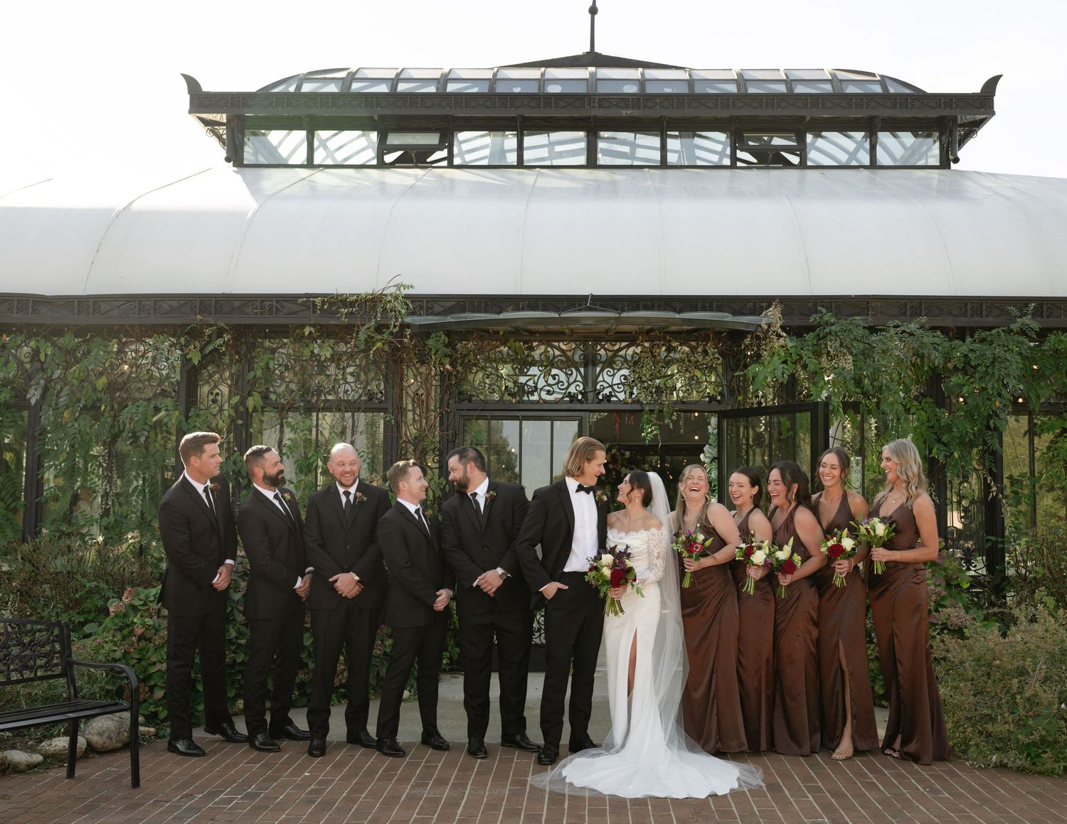 Wedding party members stand in line on a rustic wooden deck surrounded by lush greenery and trees.