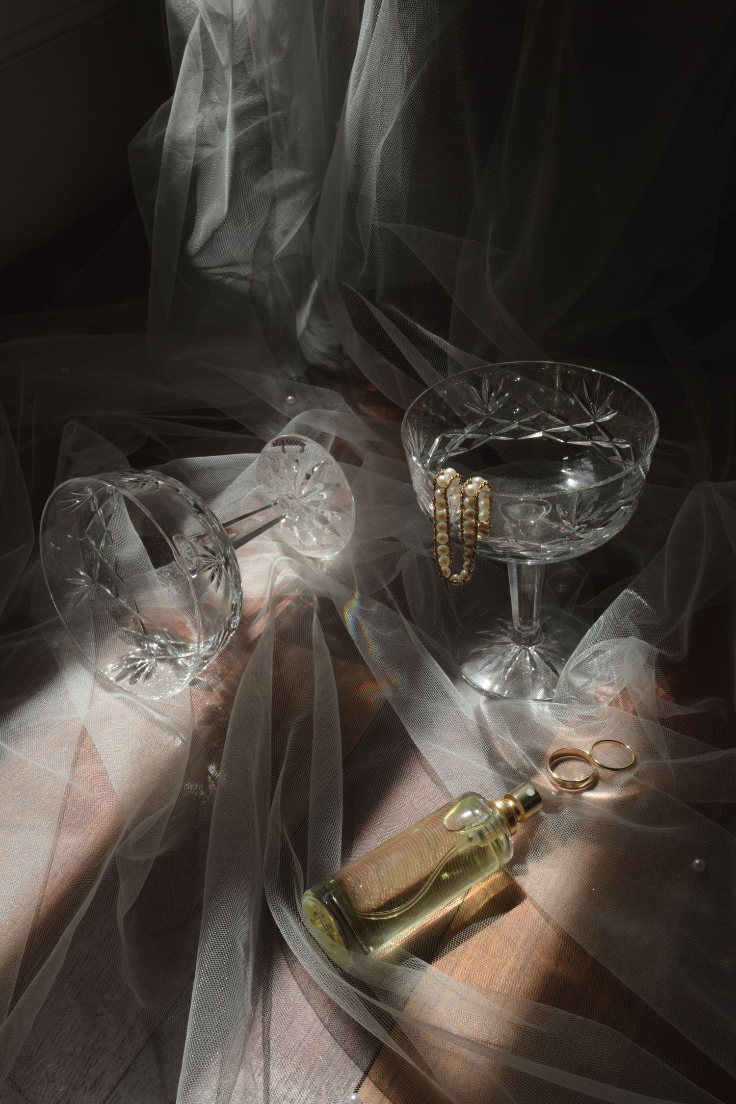Vintage crystal glasses and jewelry arranged with flowing tulle in dramatic lighting.