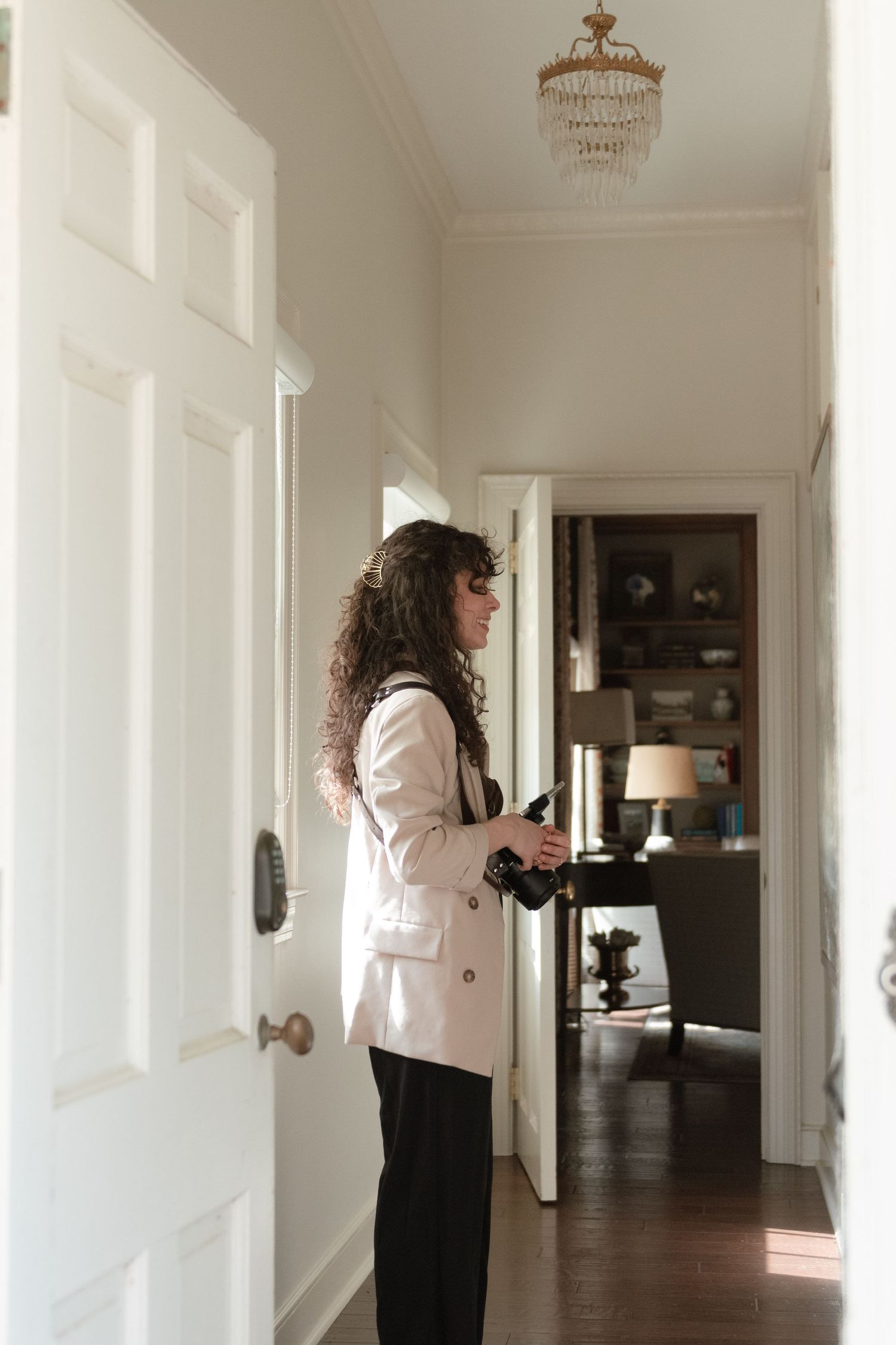 A person in a white blazer and dark pants stands in an elegant hallway with a crystal chandelier and white doors.