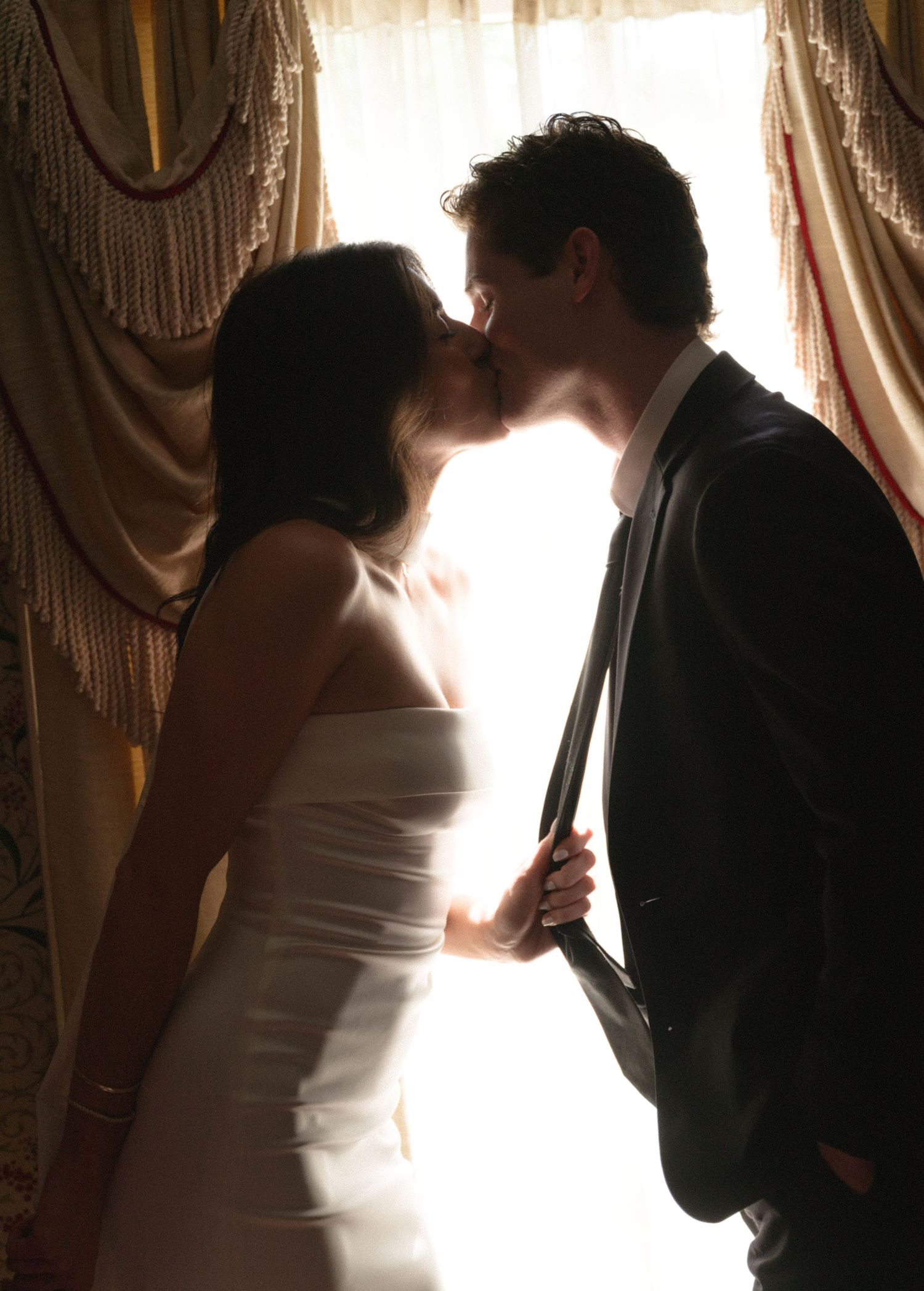 Silhouetted couple in wedding attire against backlit window with dramatic lighting.