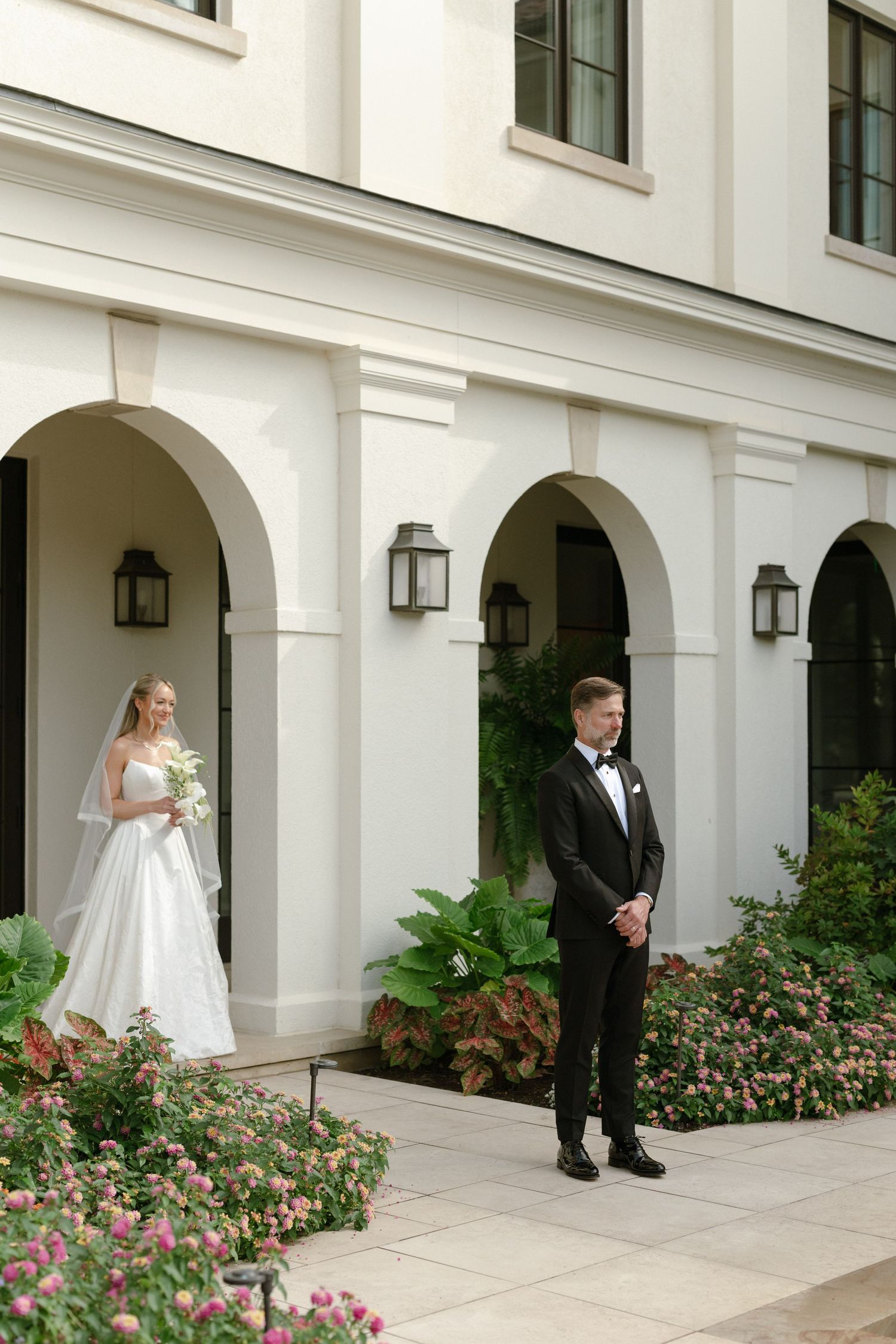 Couple in formal wedding attire pose separately in arched white hallway with pink flowers.