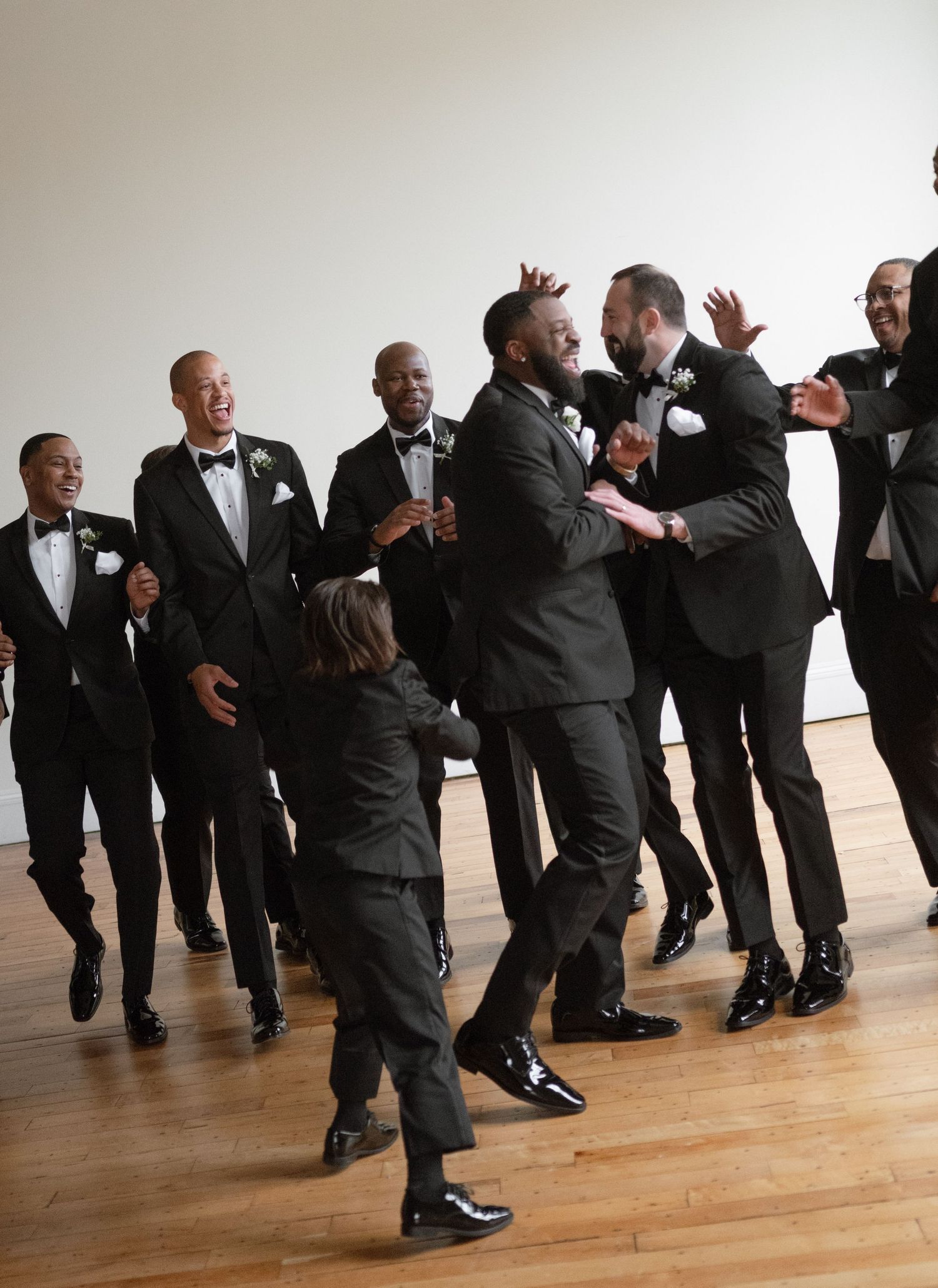 Groomsmen in black tuxedos and bowties dancing joyfully on a wooden floor.