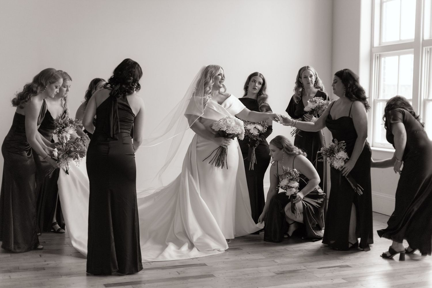 Wedding party in formal black dresses gathered around bride in elegant white gown.