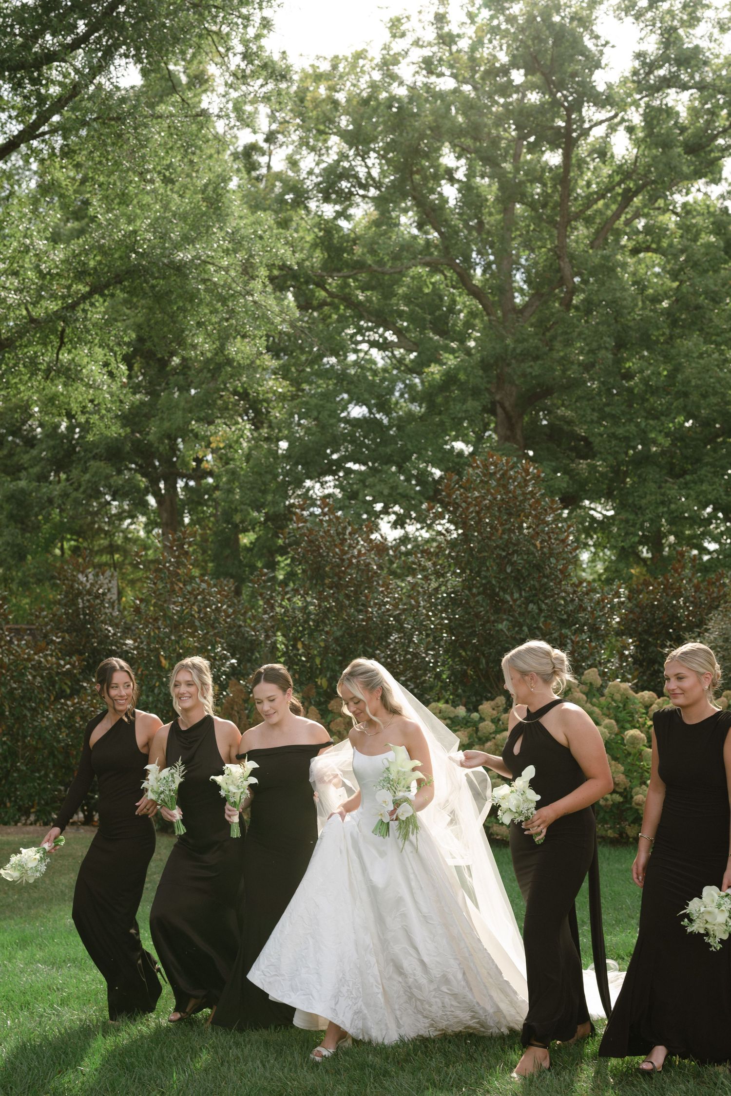 A wedding party in black dresses and white gown walks together in a line across a green lawn on a sunny day.