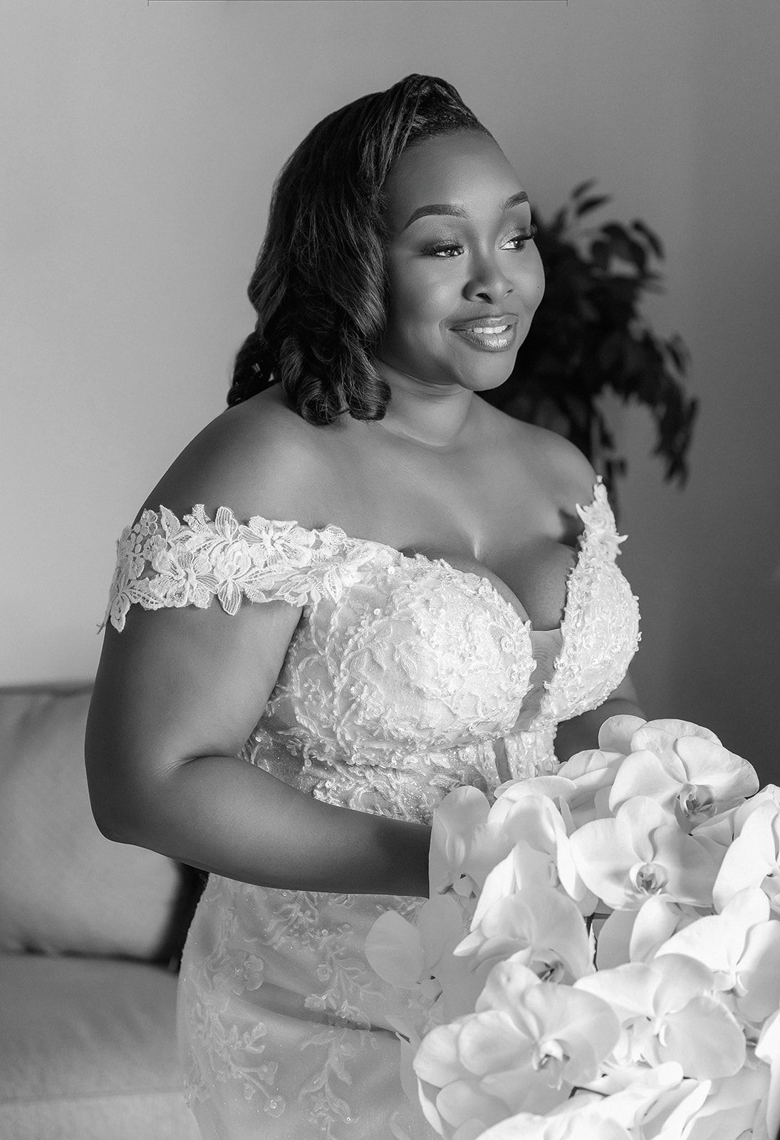 Black and white portrait of a bride in off-shoulder lace gown holding white orchid bouquet, smiling elegantly.