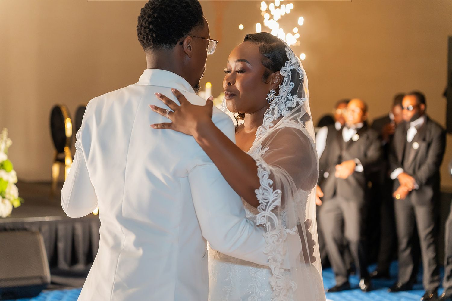 Bride and groom share their first dance, bride wearing lace veil, with sparklers and guests in background.