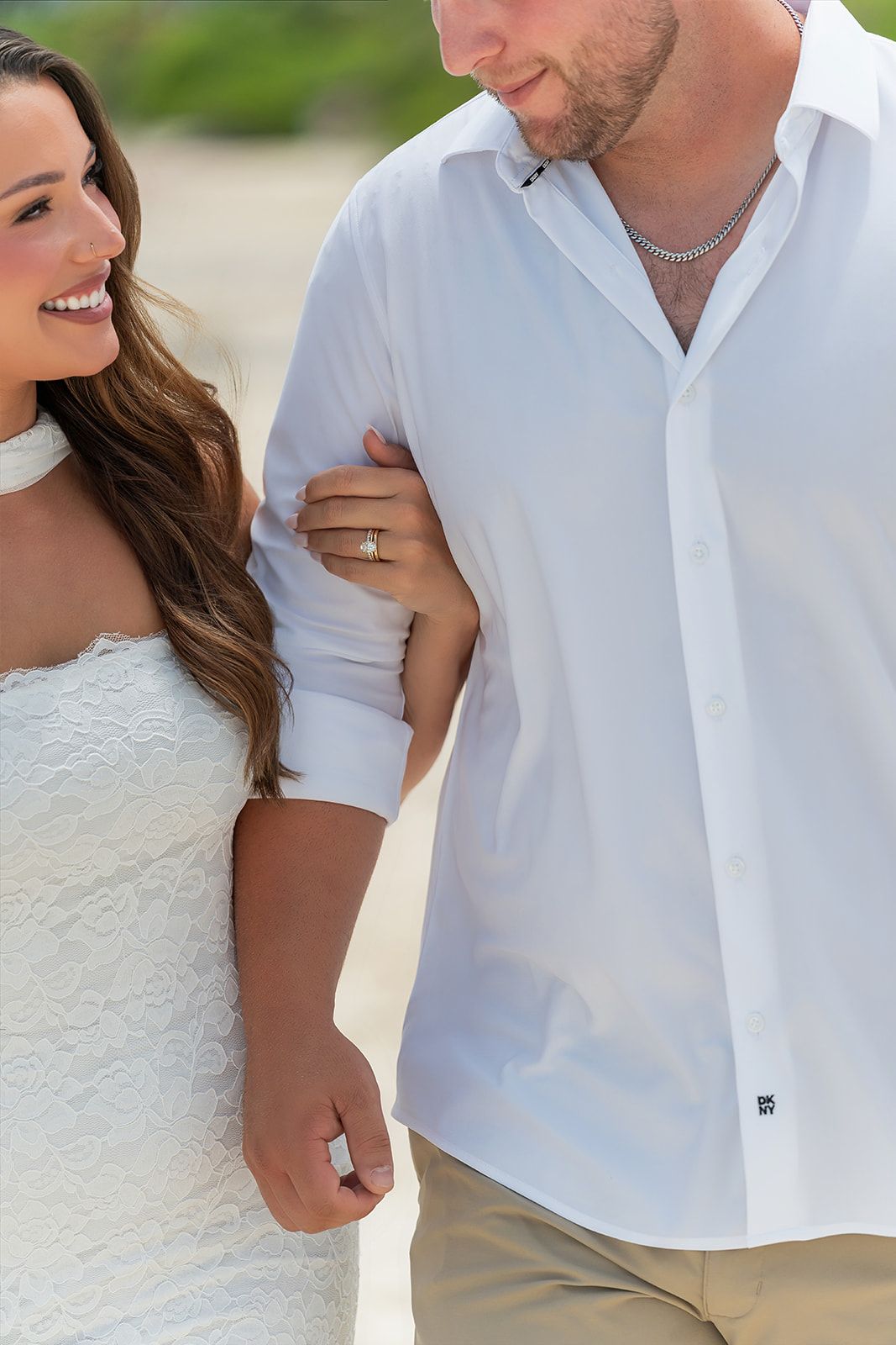 Happy couple in white outfits walking on a beach, woman showing engagement ring while holding man's arm.