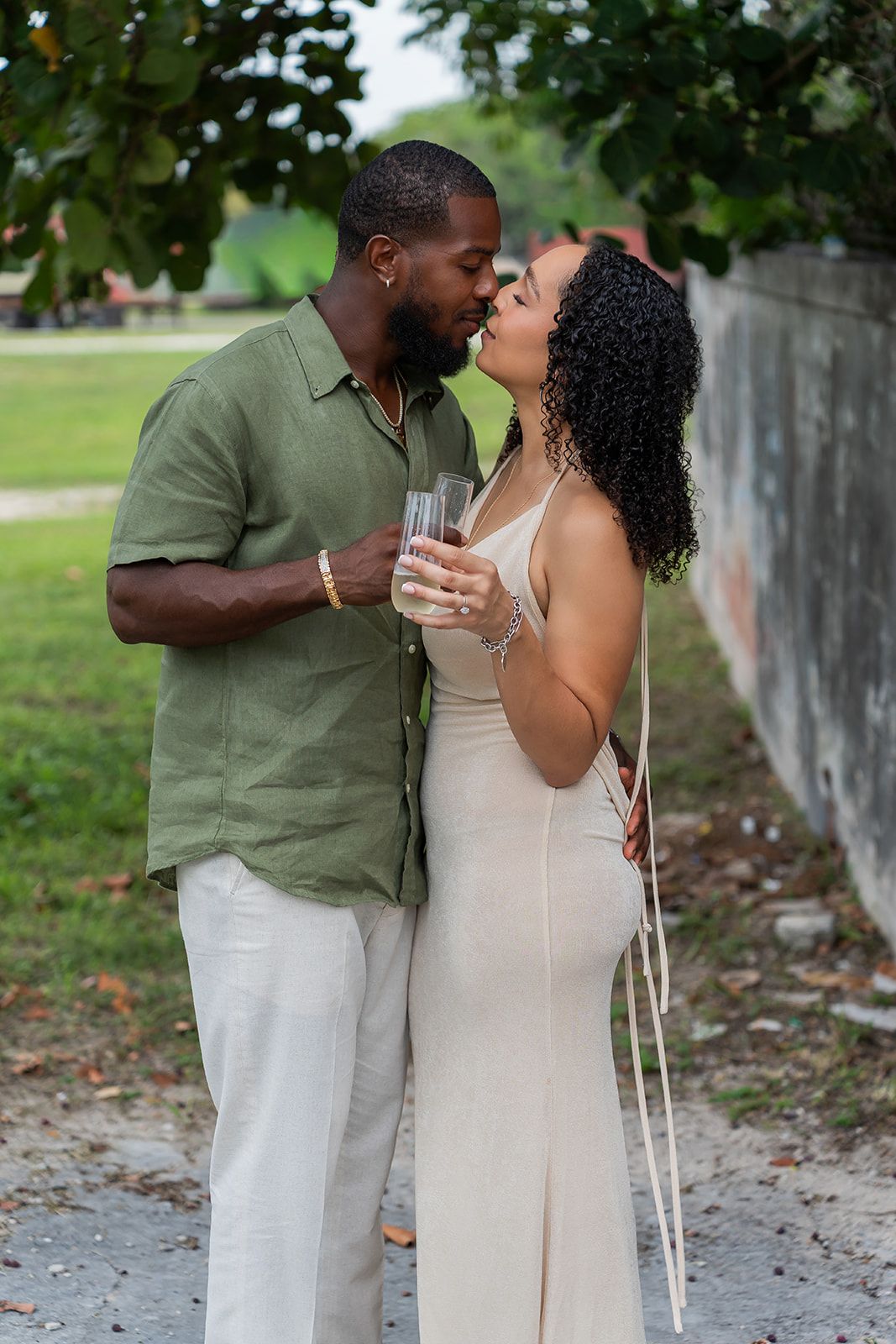 A couple shares a romantic kiss outdoors, the man in a green shirt and the woman in a cream dress holding champagne glasses.