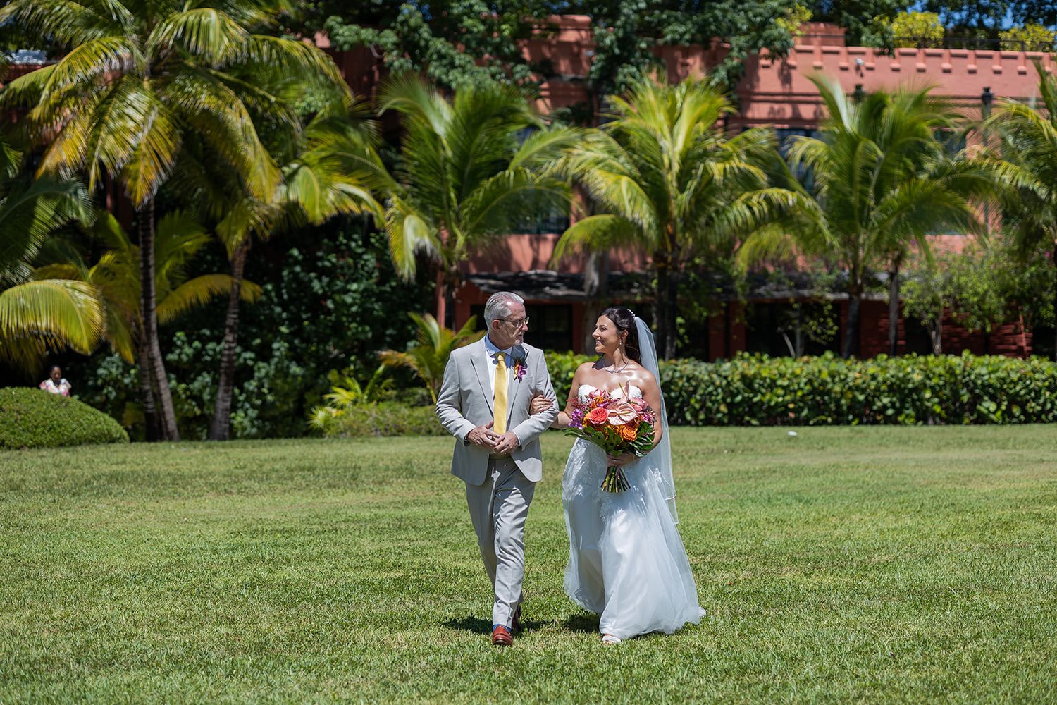 Bride and groom walking on lush green lawn surrounded by tropical palm trees at an outdoor wedding venue.